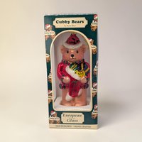 A large glass bear ornament wearing a red and white Santa hat and holding a green present. The texture is a shimmering, metallic hand-painted glass with a tall, sculpted silhouette and a gold hanging loop.