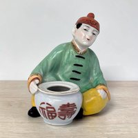A porcelain figurine of a seated person in a green robe and red cap leaning over a small white pot. The pot features red calligraphic characters, and the figure is set against a plain background on a light wood surface.