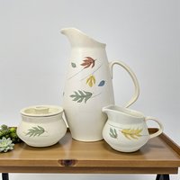 A three-piece ceramic service set featuring a tall, elegant pitcher and matching cream and sugar vessels. The set is finished in a warm tan speckled glaze and decorated with vibrant autumnal leaf motifs in shades of orange, yellow, and brown.