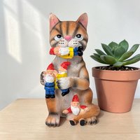 A textured resin figurine of a brown tabby cat with a pink nose, sitting upright while holding and biting several small garden gnomes with red hats. The cat has a mischievous expression and a sculpted, fur-like finish.