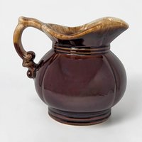A squat, round ceramic pitcher with a wide pouring spout and a large integrated handle. It features a glossy dark brown glaze with a thick, frothy cream-colored drip effect around the top rim.