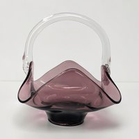 A hand-blown amethyst glass basket with a ruffled handkerchief-style rim and a clear glass arched handle. The deep purple body transitions from dark to translucent, featuring a smooth, polished texture and a pedestal base.
