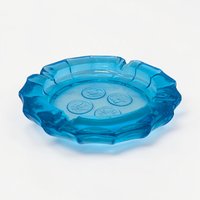 A thick, translucent ice blue glass ashtray with a scalloped edge. Four circular medallions featuring frosted patriotic "coin" motifs, including an eagle and a torch, are embossed into the center of the heavy glass base.