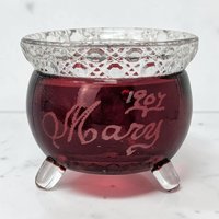 A small, three-footed glass kettle with a deep ruby red body and clear, diamond-cut rim. The red surface features an etched cursive name, Mary, and the year 1907. The texture is smooth glass contrasted by the textured, geometric collar.