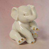A small, seated ivory porcelain elephant with its trunk raised. The figurine features a smooth, creamy glaze with gold-gilded accents on the toes. It has a gentle, peaceful expression.