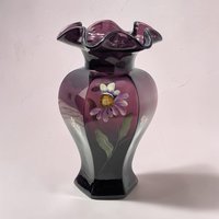 A tall, elegant vase in deep mulberry purple glass with a satin finish. It features hand-painted white floral sprigs and soft green leaves. The vase has a flared, ruffled rim and a rounded body.