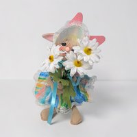 Tan felt bunny wearing a colorful striped sun hat and matching dress with white lace trim. She holds a large bouquet of three white daisies with yellow centers. Her tall ears are lined in pink felt and her face has a cheerful hand-painted smile.