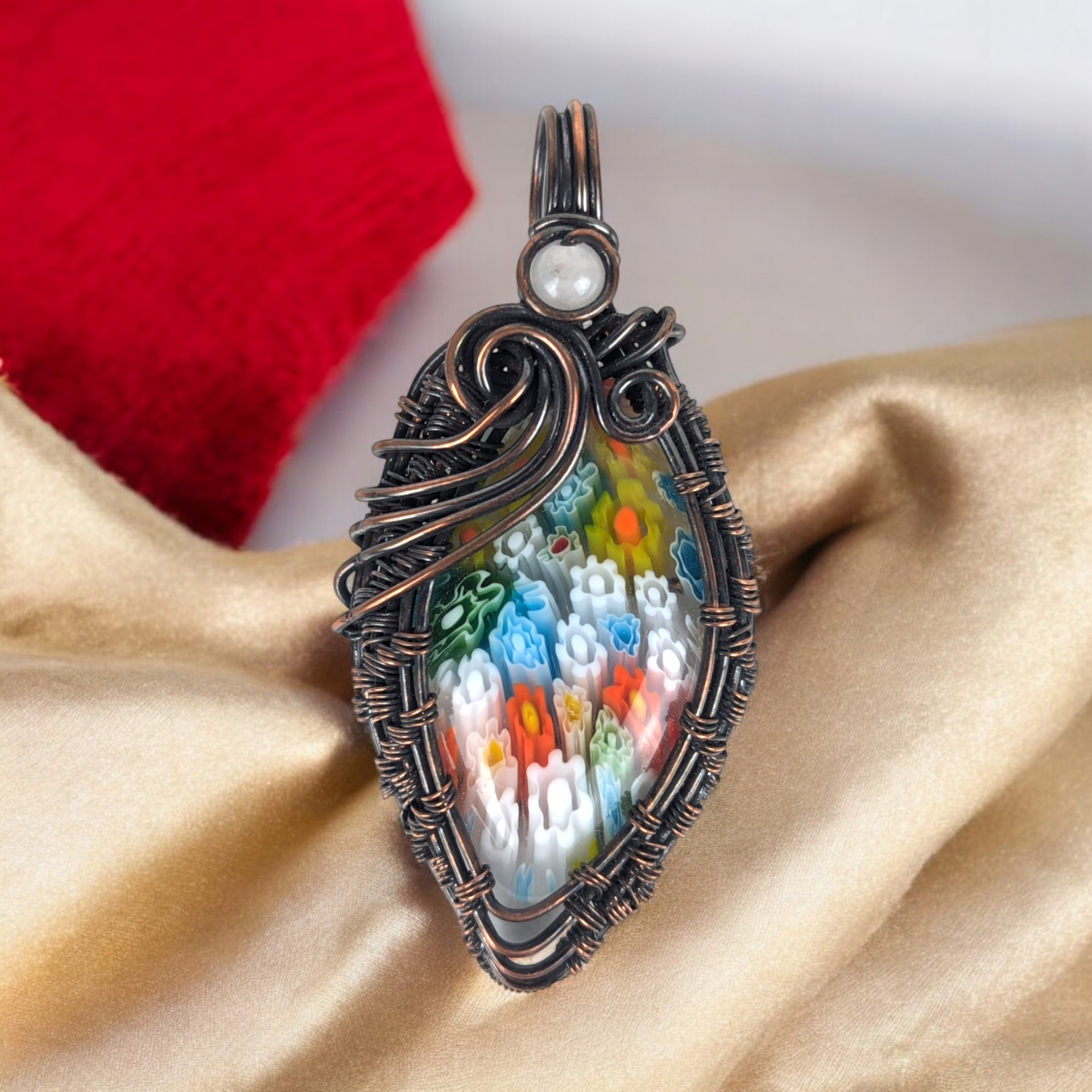 An oval glass millefiori cabochon with a colorful mosaic of tiny floral patterns in red, blue, and yellow. It is encased in oxidized copper wire with a small pink rose quartz bead nestled into the wirework above the stone.