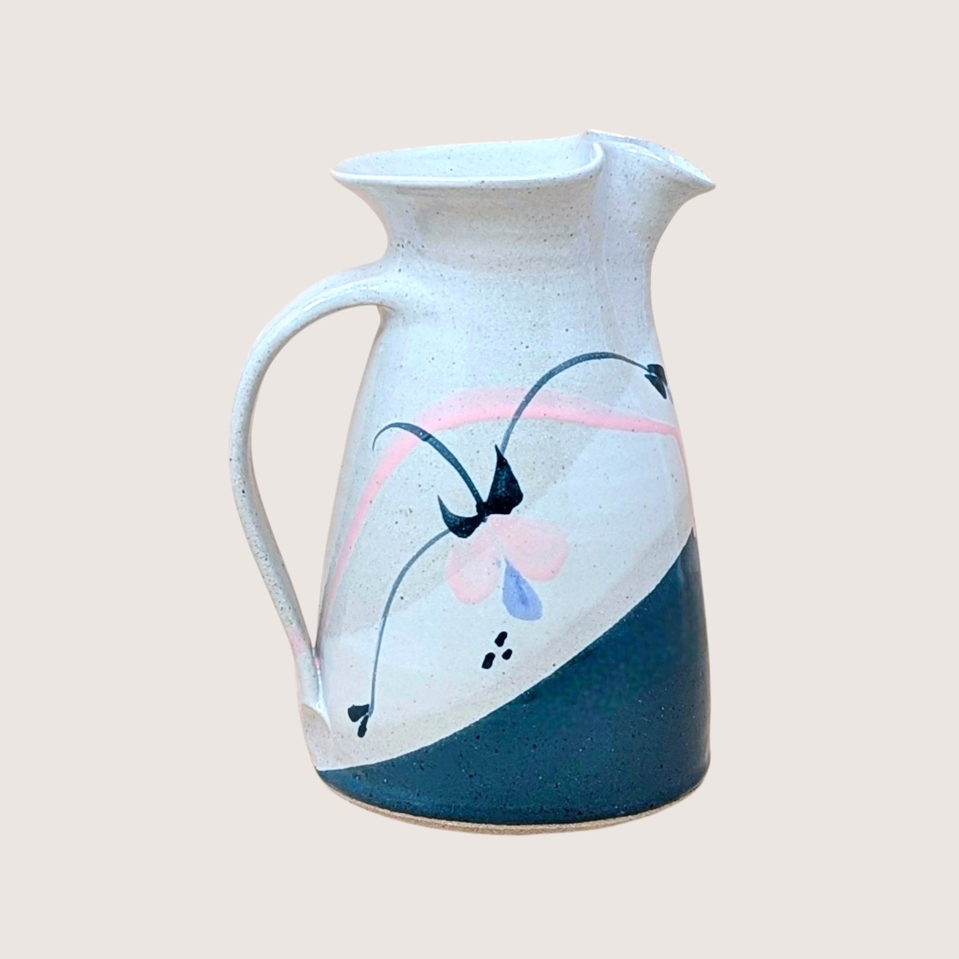 Speckled cream earthenware pitcher with a wide flared rim and sturdy handle. Features a hand-painted abstract floral design in teal, pink, and indigo with a diagonal teal color-block base.