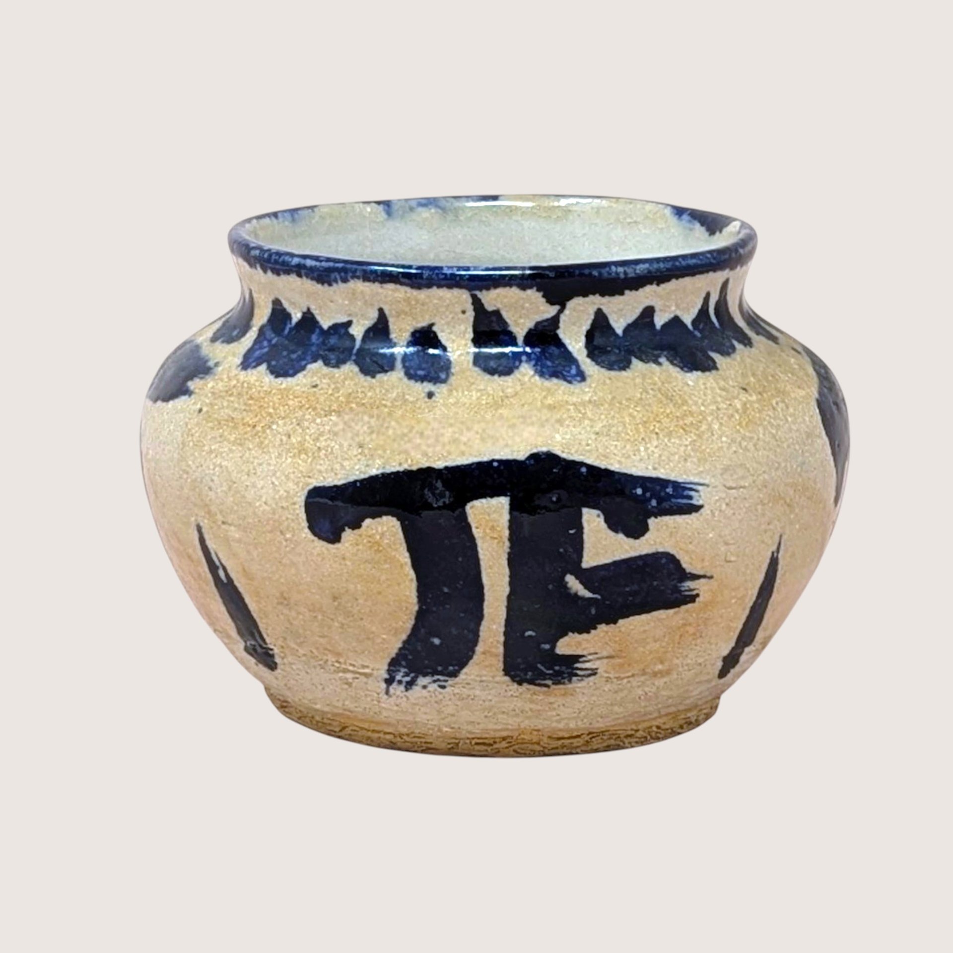 Hand-thrown pottery bowl with a globular shape and a sandy, textured tan glaze. Featuring bold, gestural cobalt blue brushstrokes including a large "PI" or "TF" symbol and a saw-tooth rim pattern.