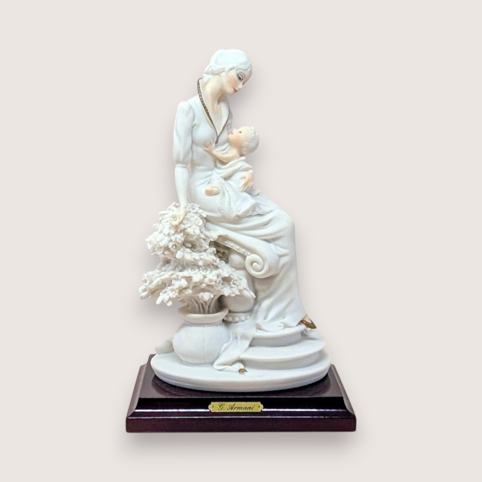 Giuseppe Armani "Maternal" porcelain figurine of a mother cradling an infant, seated beside a flowering tree. Set on a dark wood square pedestal base with a gold nameplate. Matte white and cream finish.