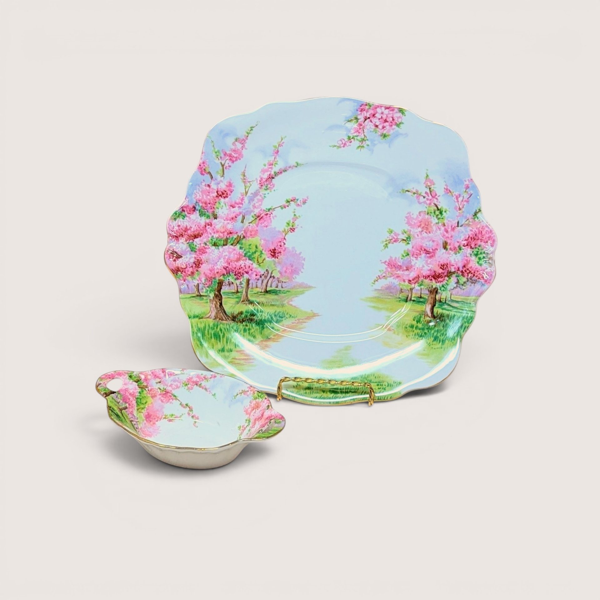 Two-piece Royal Albert Blossom Time bone china set. Includes a 9.75-inch square handled cake plate and a 5.5-inch oval handled sweet meat dish. Features pink blossom trees and gold-trimmed scalloped edges.