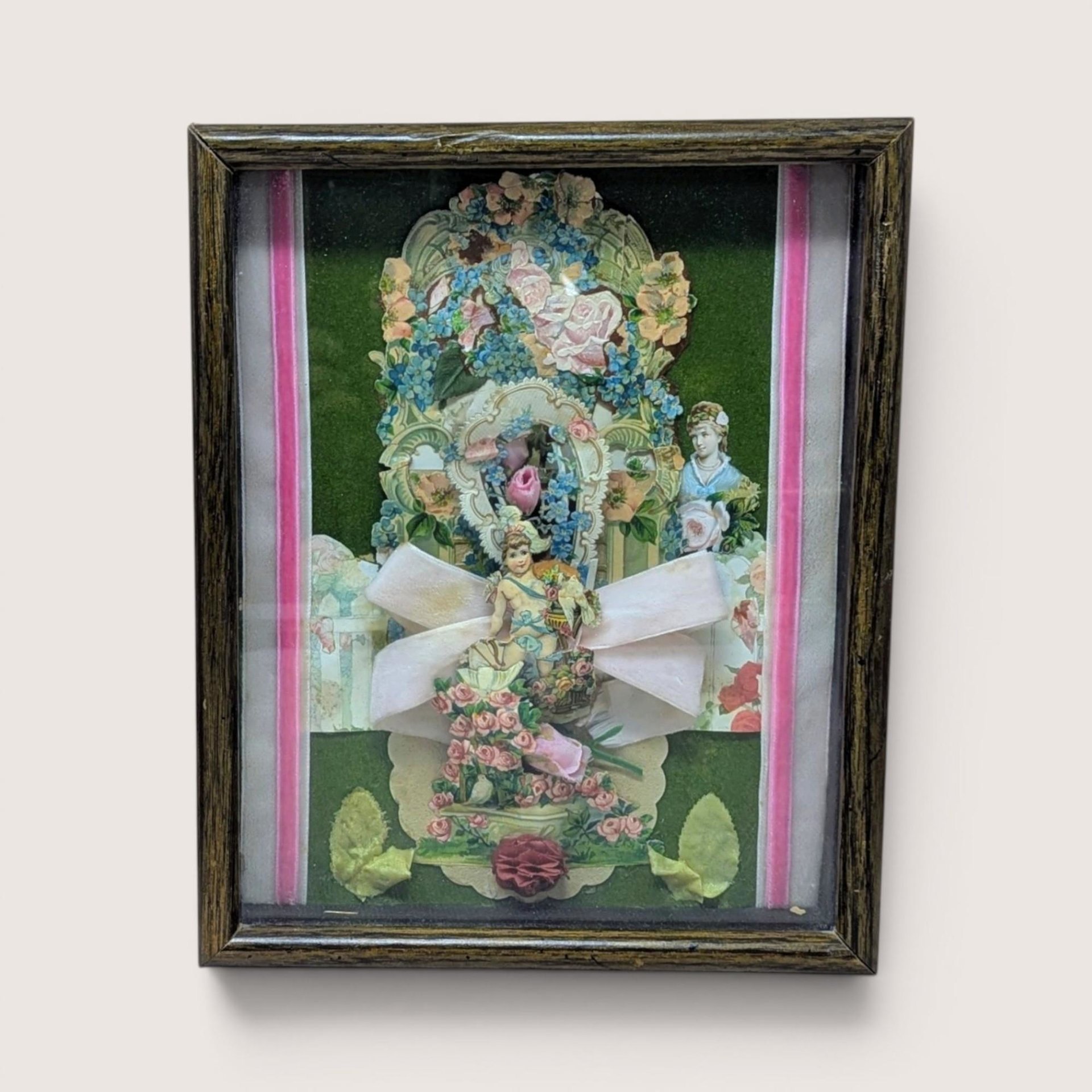 A vertical shadow box frame containing a Victorian-style paper collage. Features a cherub, floral lithographs, and a large pink velvet bow against a forest green velvet background. Housed in a dark wood-grain frame.
