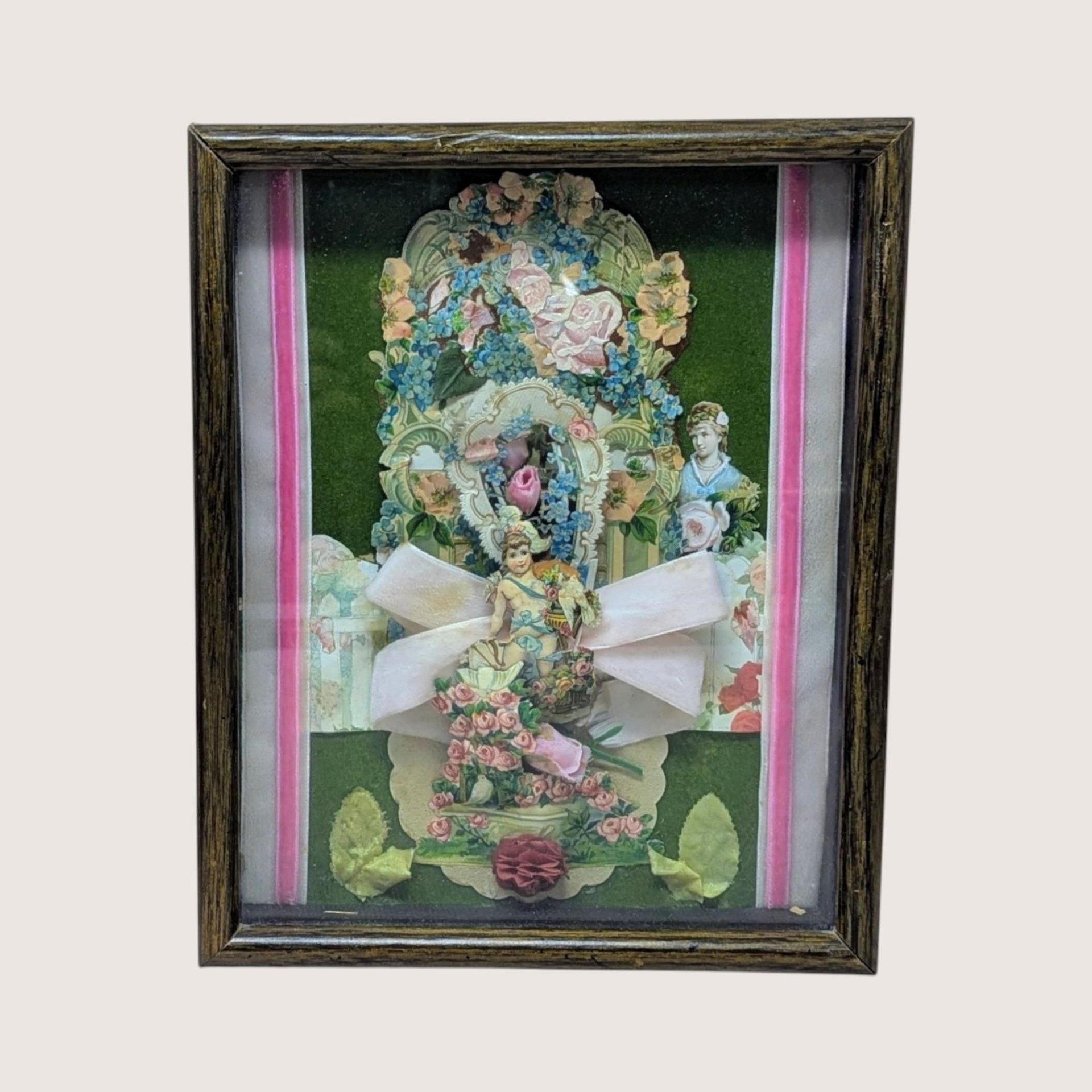A vertical shadow box frame containing a Victorian-style paper collage. Features a cherub, floral lithographs, and a large pink velvet bow against a forest green velvet background. Housed in a dark wood-grain frame.