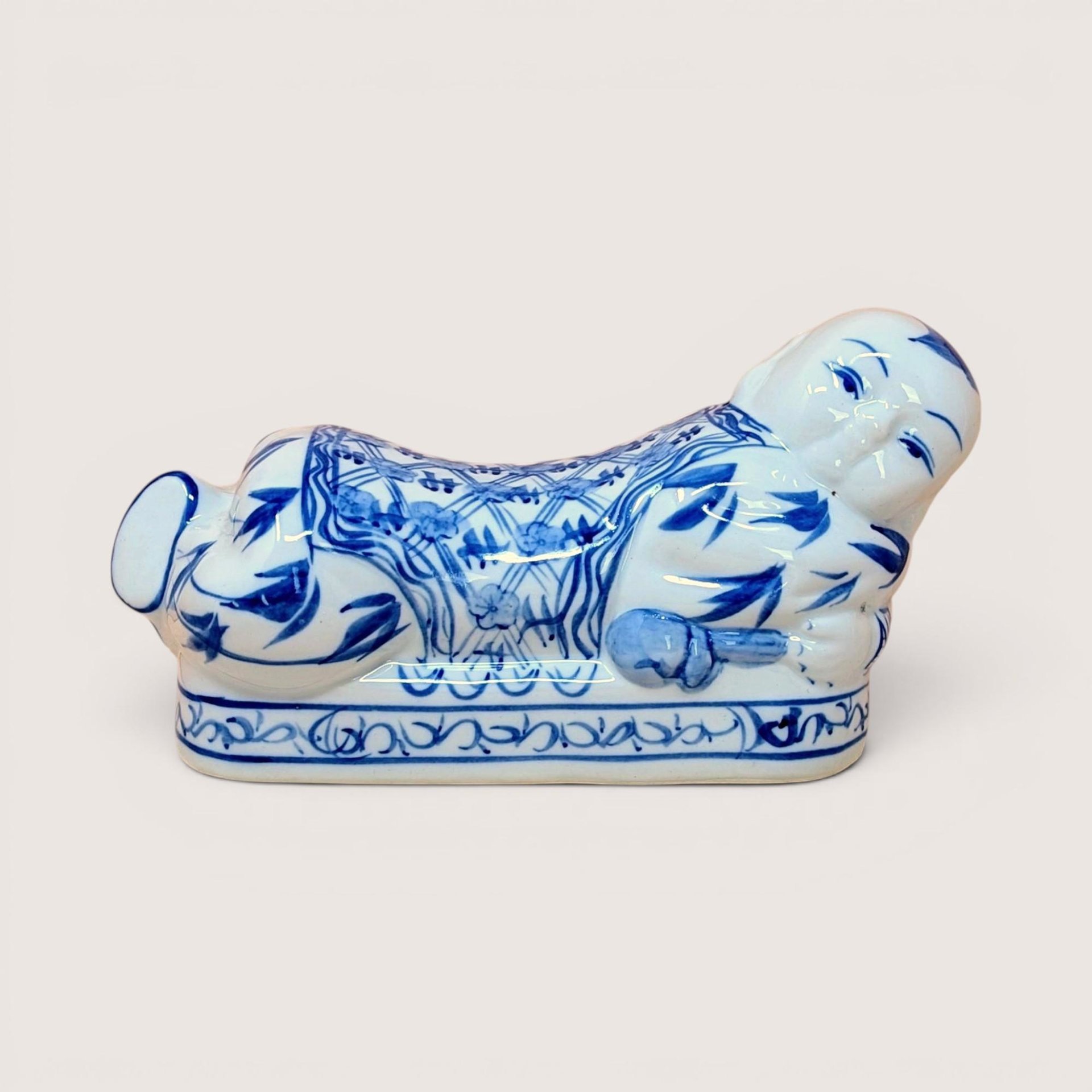 A horizontal porcelain headrest shaped like a reclining child. Featuring a crisp white ground with traditional cobalt blue floral and scroll patterns. Glossy vitrified texture with hand-painted facial details.