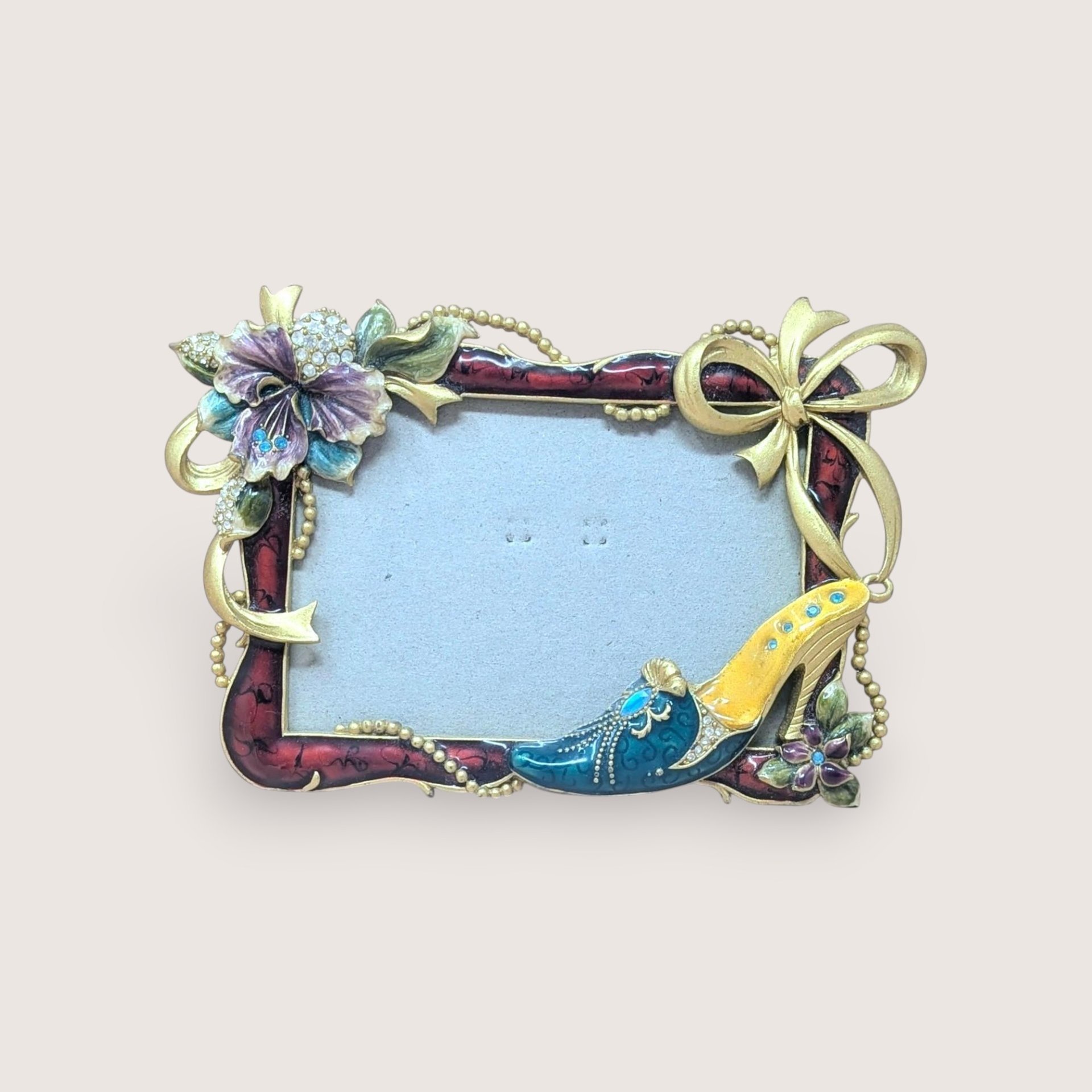 A horizontal photo frame with a deep red enamel border. Ornate gold-tone decorations include a crystal-studded purple hibiscus-style flower on the top left and a fashionable stiletto shoe on the bottom right.
