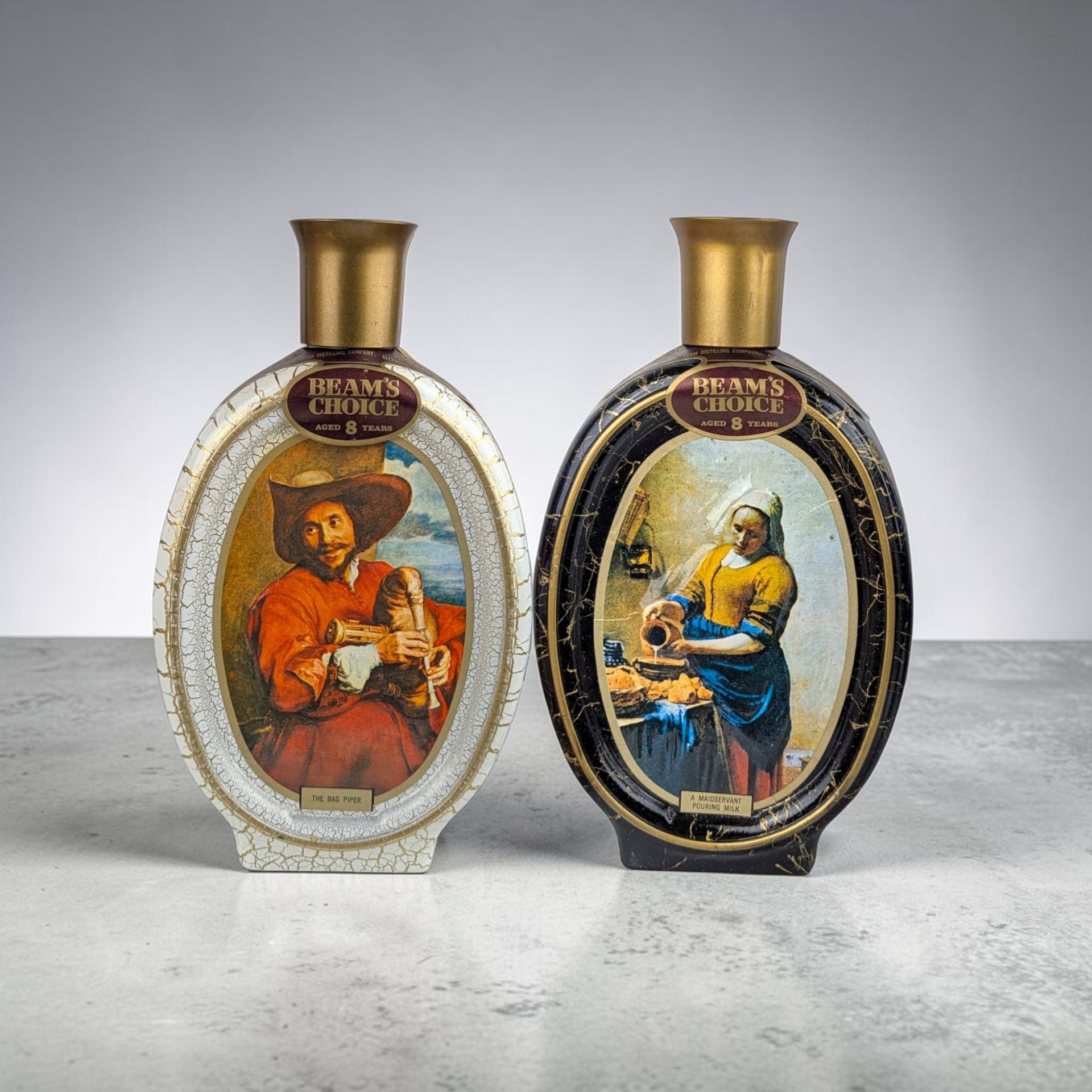 Two oval ceramic decanters. One is white with a painting of a man playing bagpipes; the other is black with Vermeer's Milkmaid painting. Both have gold flared tops.