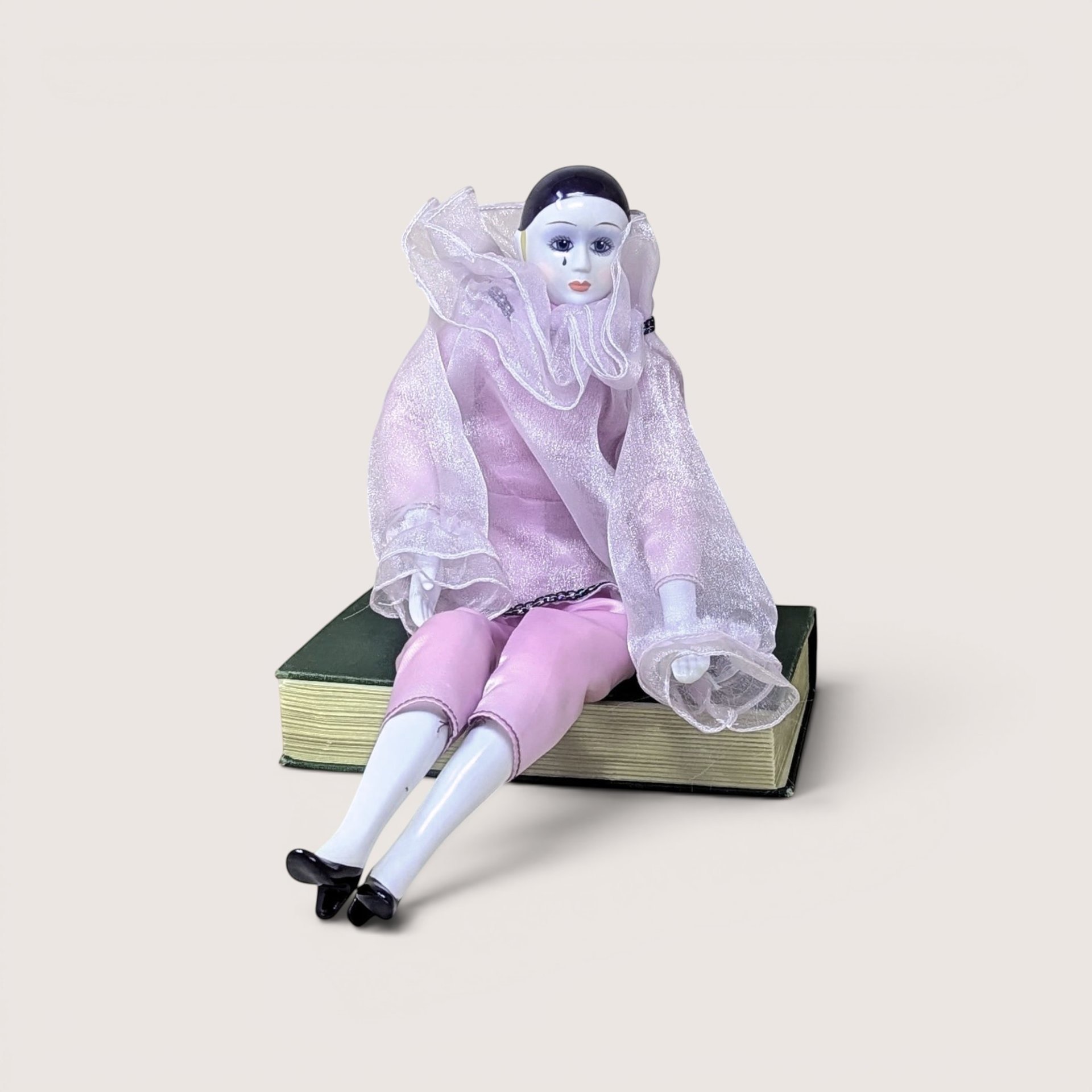 Vintage 1980s porcelain Pierrot mime clown doll dressed in a light pink fabric outfit while sitting on a green book.