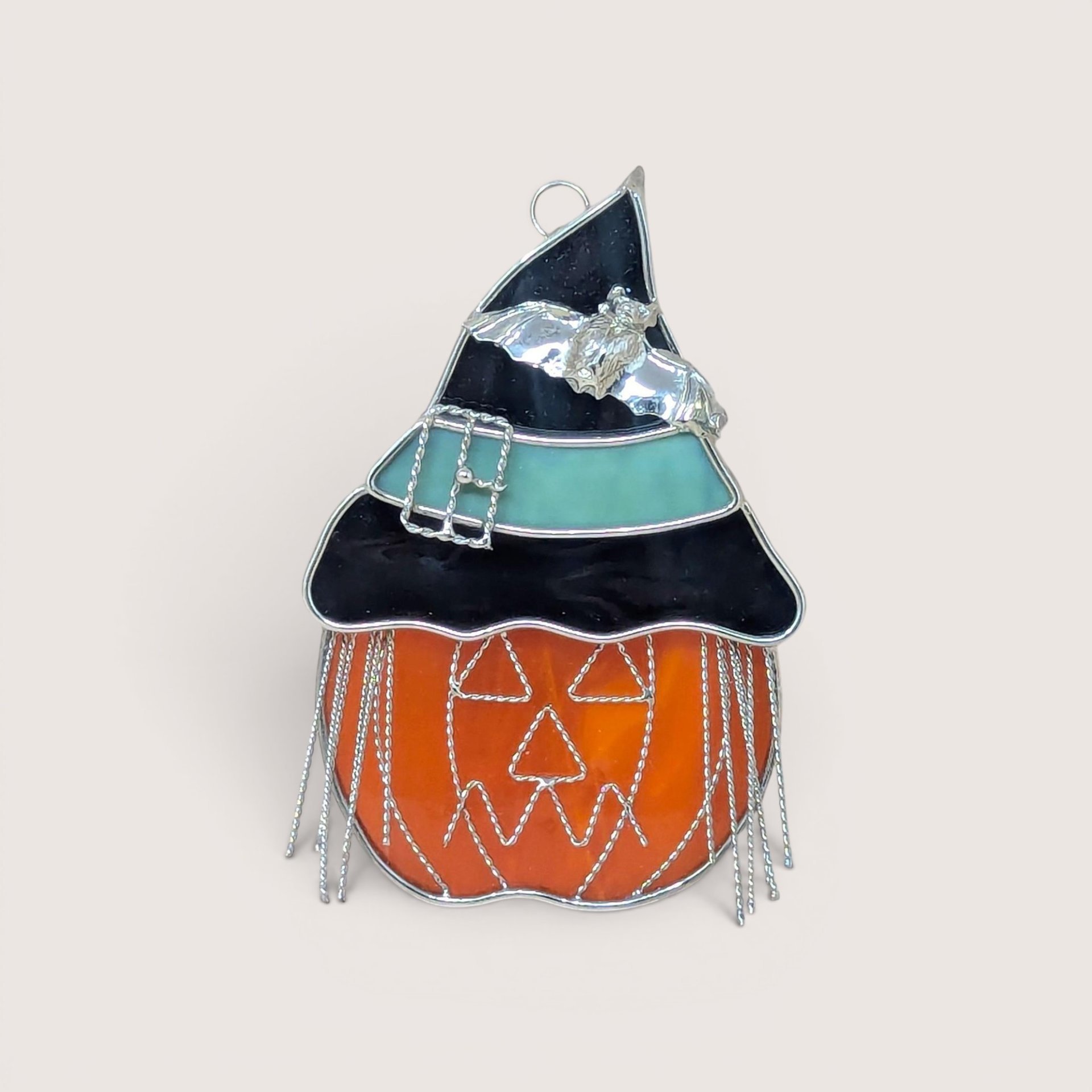 A vibrant stained glass suncatcher of a jack o' lantern wearing a black and teal witch's hat with a silver bat and wire fringe on a neutral background.