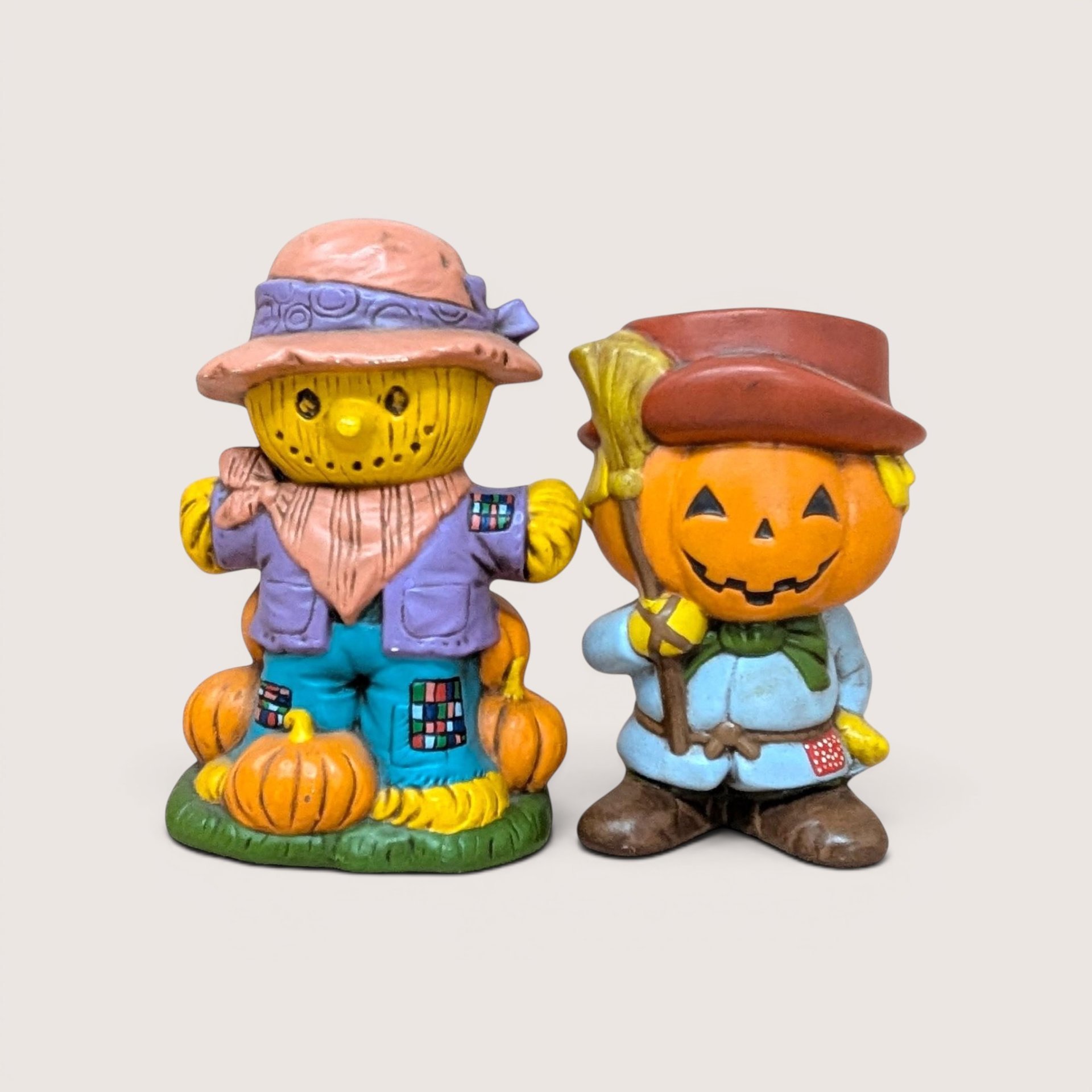 A pair of vintage hand-painted ceramic figurines featuring a friendly scarecrow standing on pumpkins and a jack o' lantern man holding a broom.