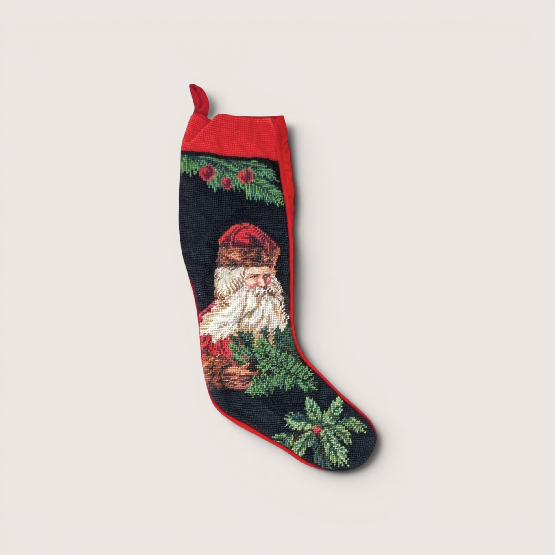 A vintage needlepoint Christmas stocking featuring a detailed Old World Santa with a fur hat and evergreen branches against a dark background.
