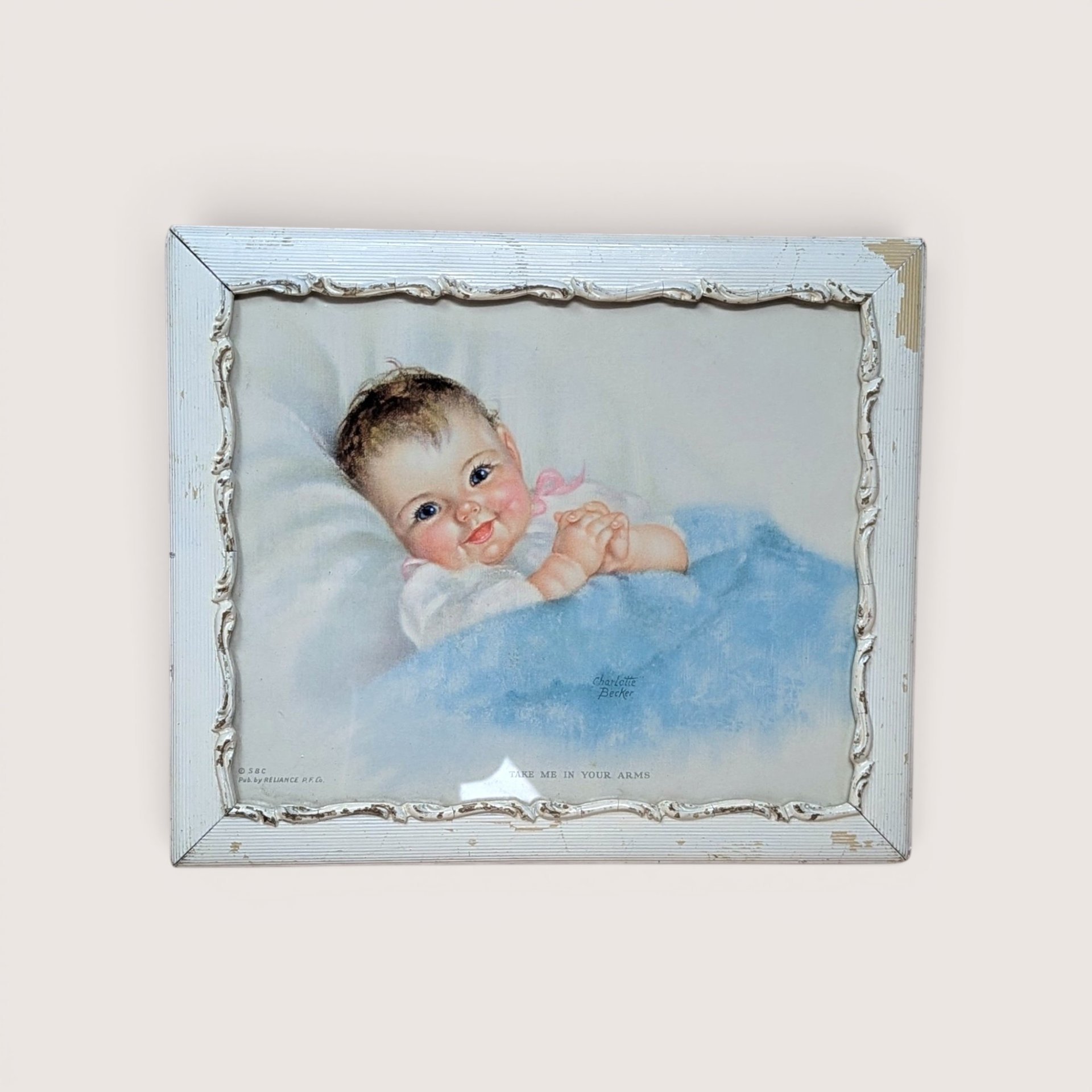 A 1940s vintage Charlotte Becker framed nursery print on a soft pink background. The sweet lithograph depicts a rosy-cheeked baby tucked under a blue blanket, housed in a distressed white decorative wooden frame with chipped paint accents.