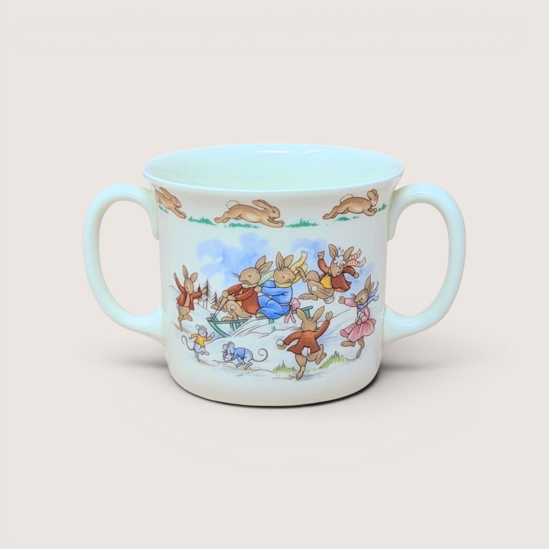 A white porcelain two-handled child's cup with illustrations of bunnies sledding, centered on a beige background.
