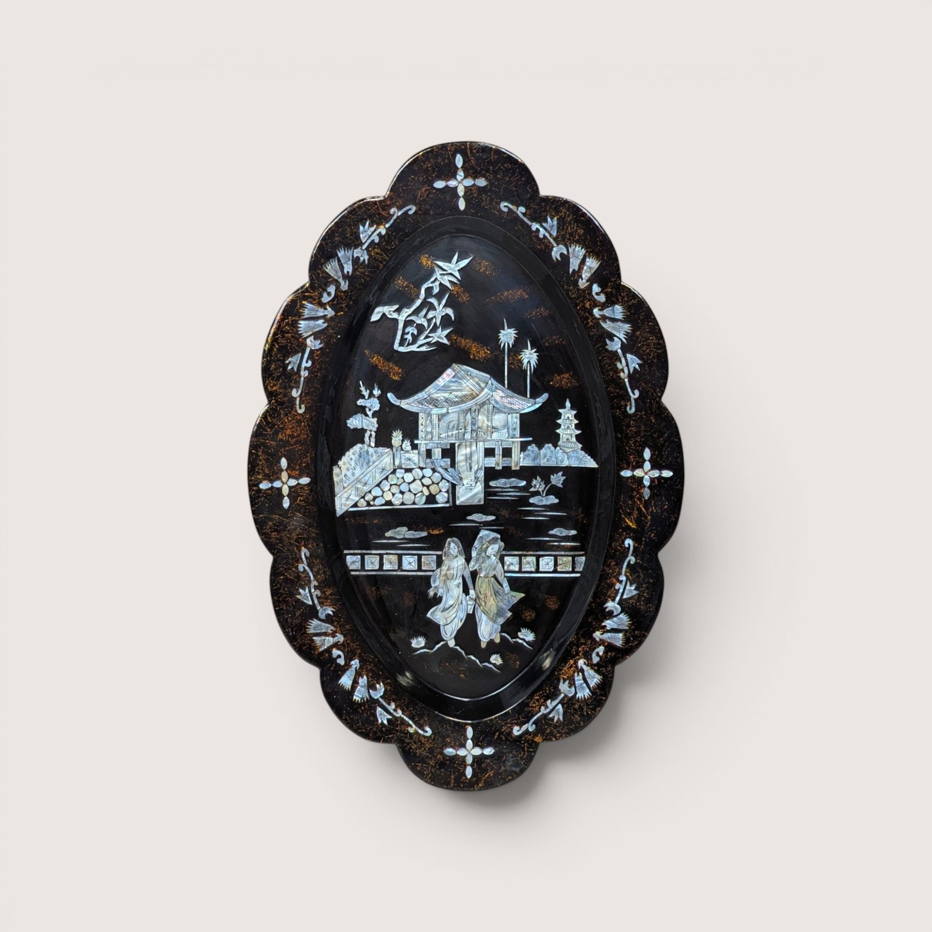 An oval black lacquer tray with mother-of-pearl inlay depicting an Asian landscape, centered on a beige background.