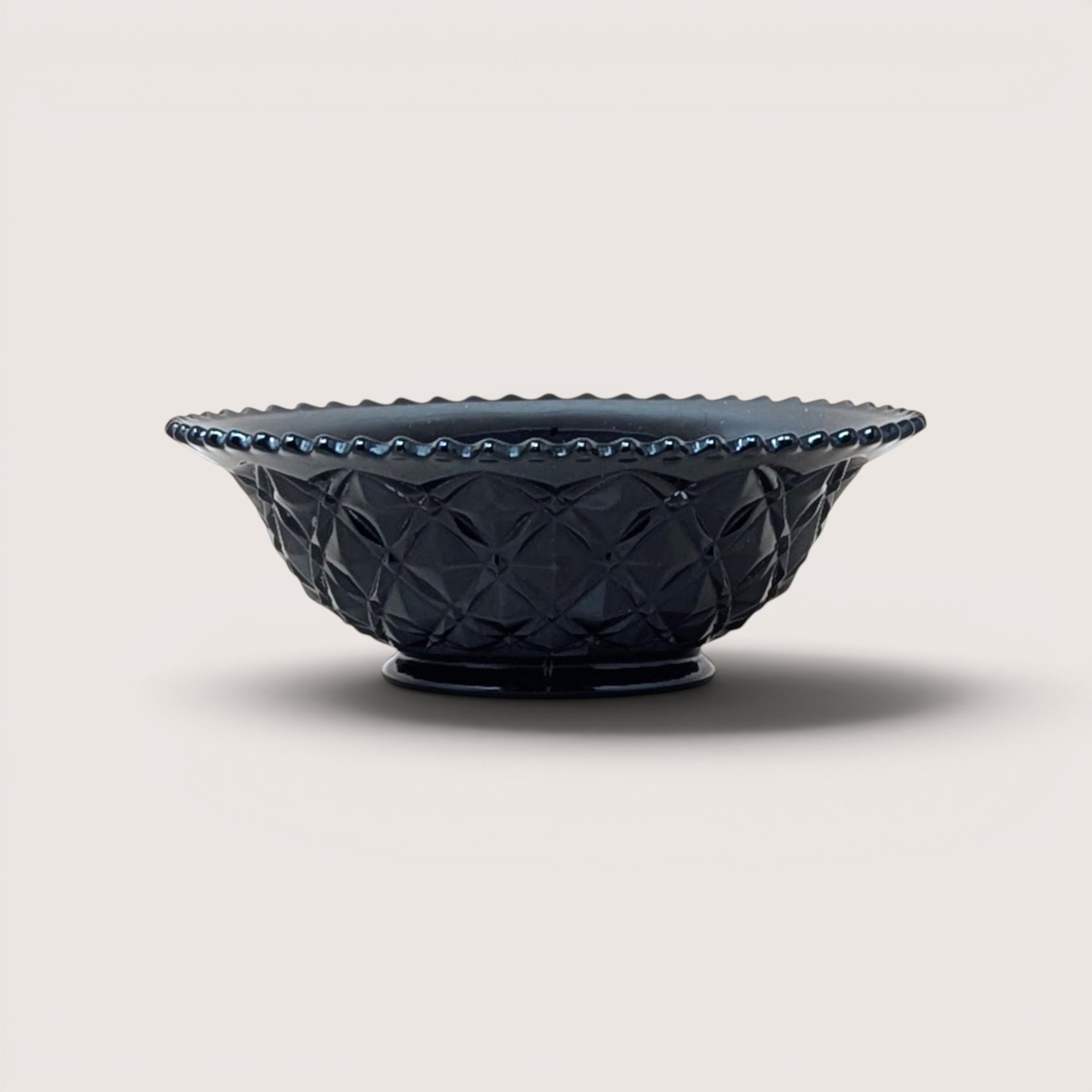 A vintage black glass bowl with a diamond pattern and beaded rim, centered on a beige background.