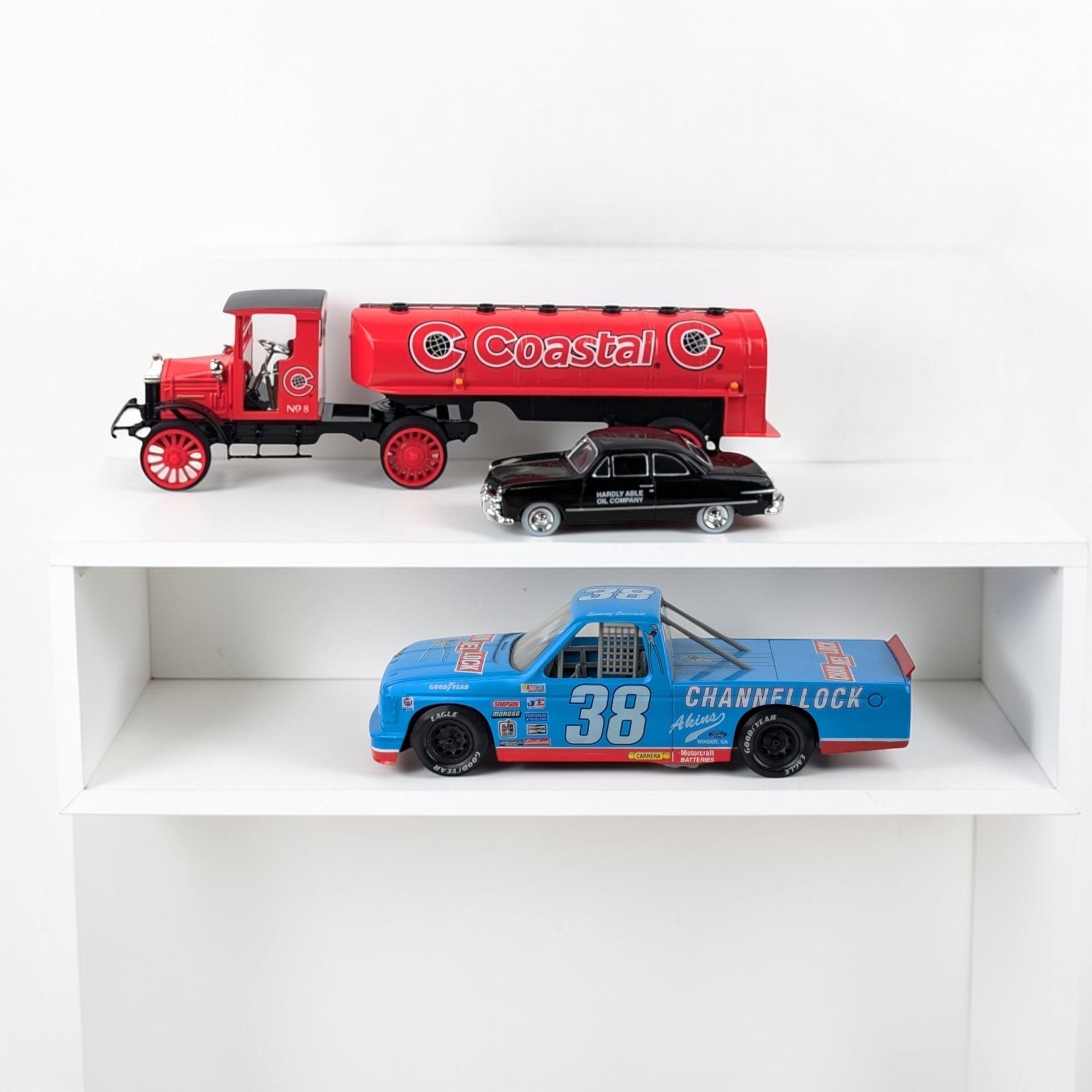 A collection of three die-cast toy vehicles displayed on white shelving. The set includes a large red Coastal tanker truck with a black cab and spoked wheels, a small black vintage sedan labeled "Hardly Able Oil Company," and a blue number 38 Channellock racing truck with various sponsor decals and a rear roll cage.