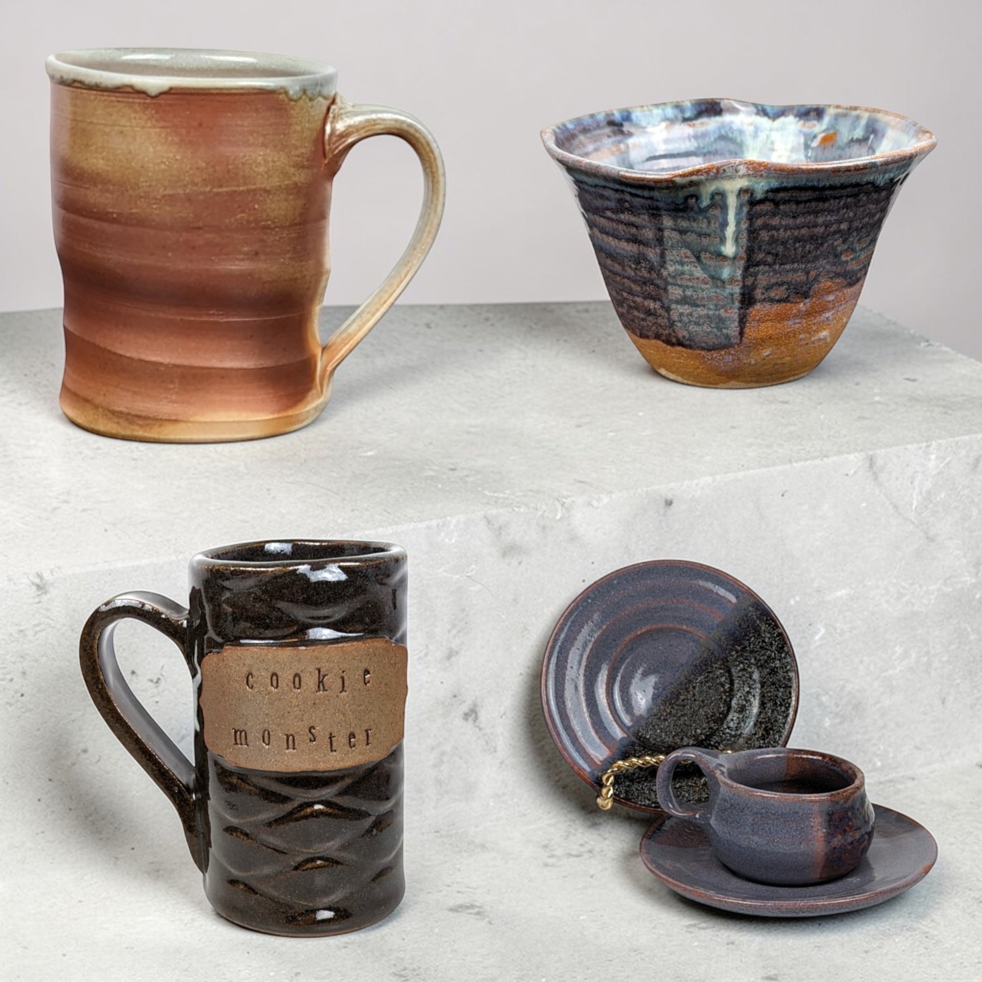 A grid of four stoneware pottery items featuring various glaze techniques. Top left is a tall cylindrical mug with a warm terracotta and tan matte finish. Top right is a flared bowl with a deep indigo blue and iron-brown drip glaze. Bottom left is a dark chocolate brown textured mug with a tan clay plaque reading "cookie monster." Bottom right is a petite dark blue cup with two matching saucers showing a subtle metallic sheen. All pieces exhibit a rustic, hand-thrown silhouette.