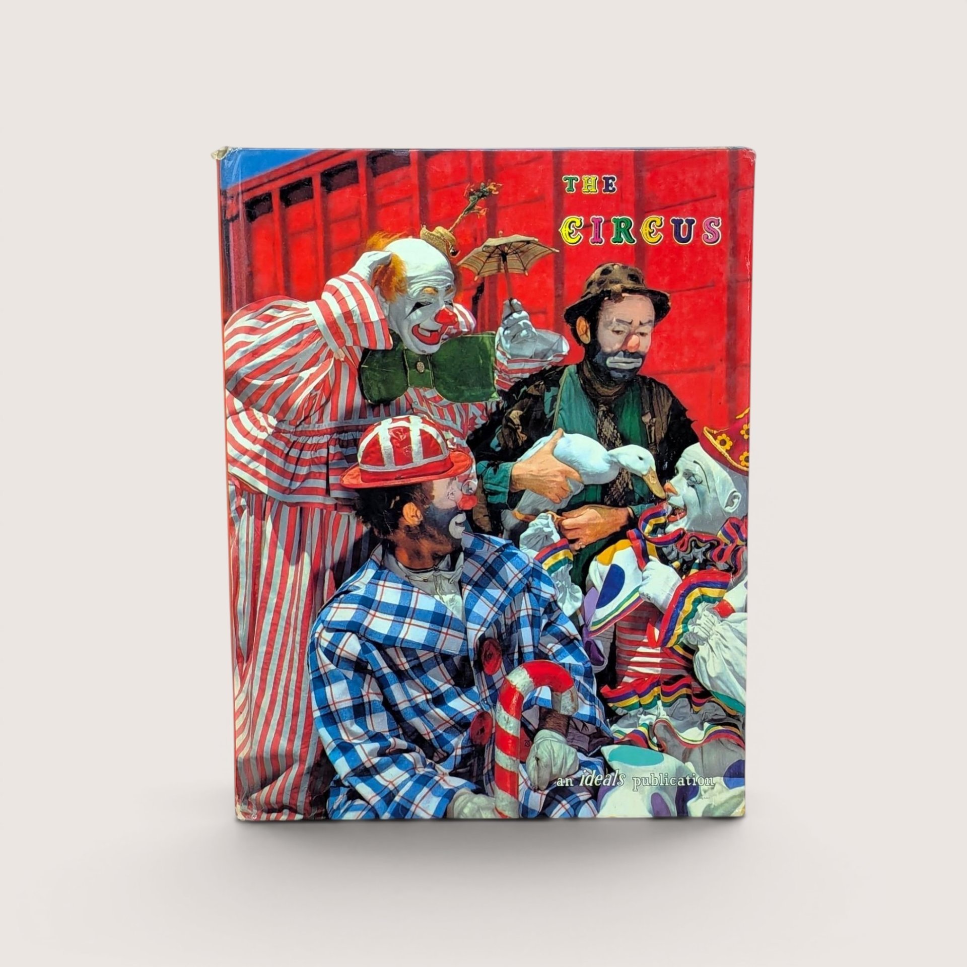 A 1961 vintage Ideals Publication book titled The Circus on a Dune colored background. The hardcover front features a vibrant red illustration of mid-century circus clowns in colorful striped and plaid costumes gathered together with a white duck.
