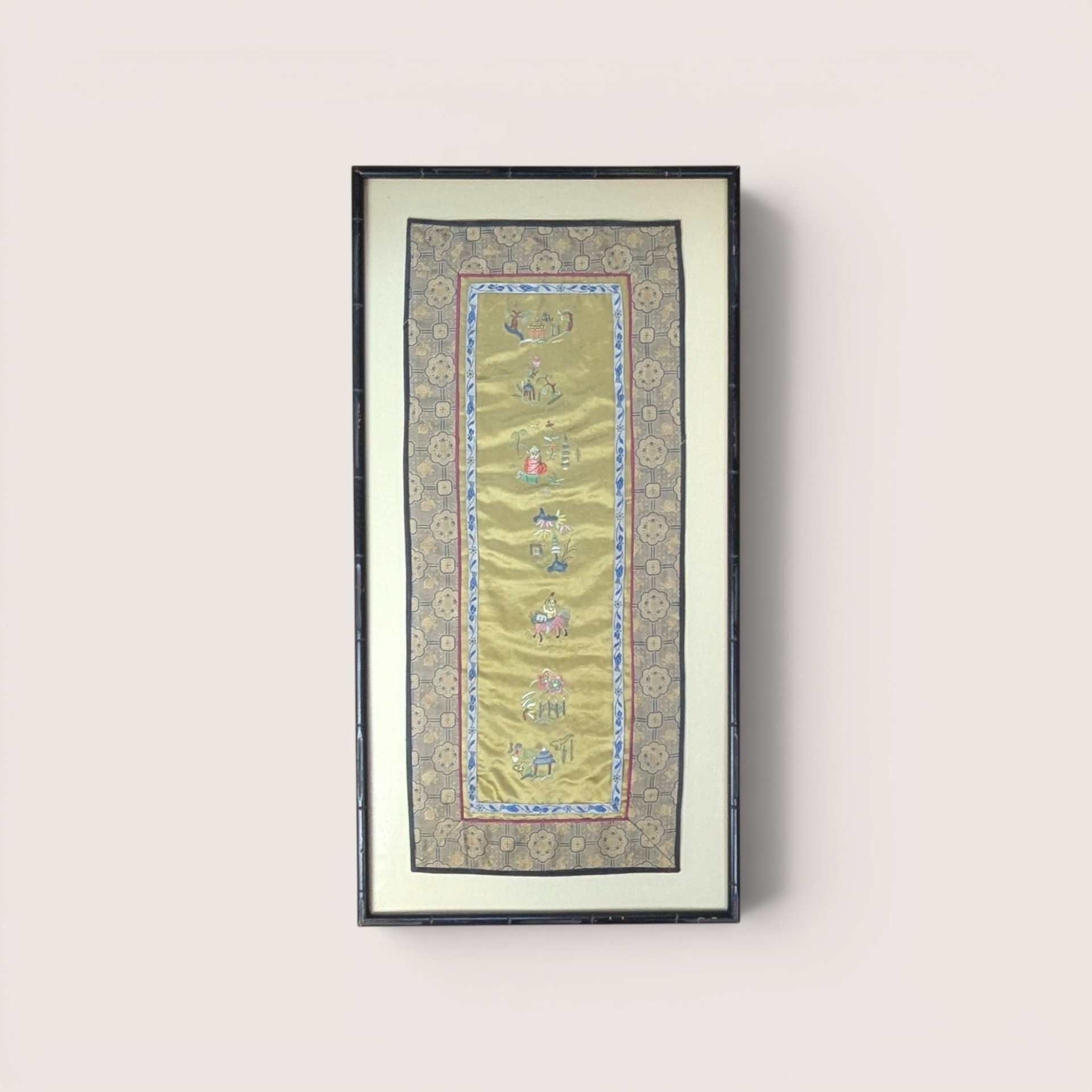 A framed vertical Chinese silk embroidery textile with small figures and decorative borders, on a beige background.