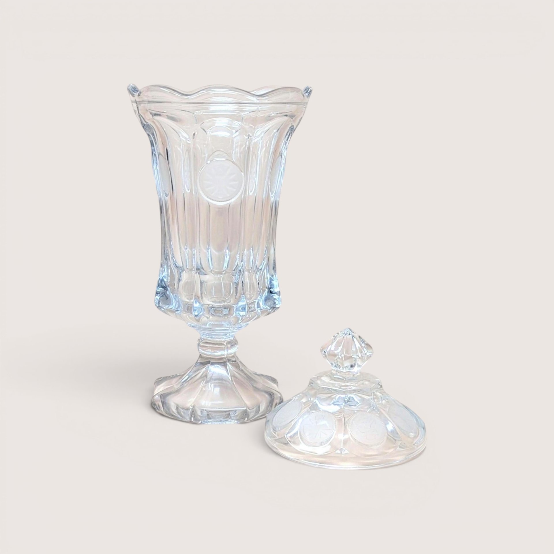 A clear glass footed urn jar with a ruffled rim next to its matching lid featuring frosted coin motifs.