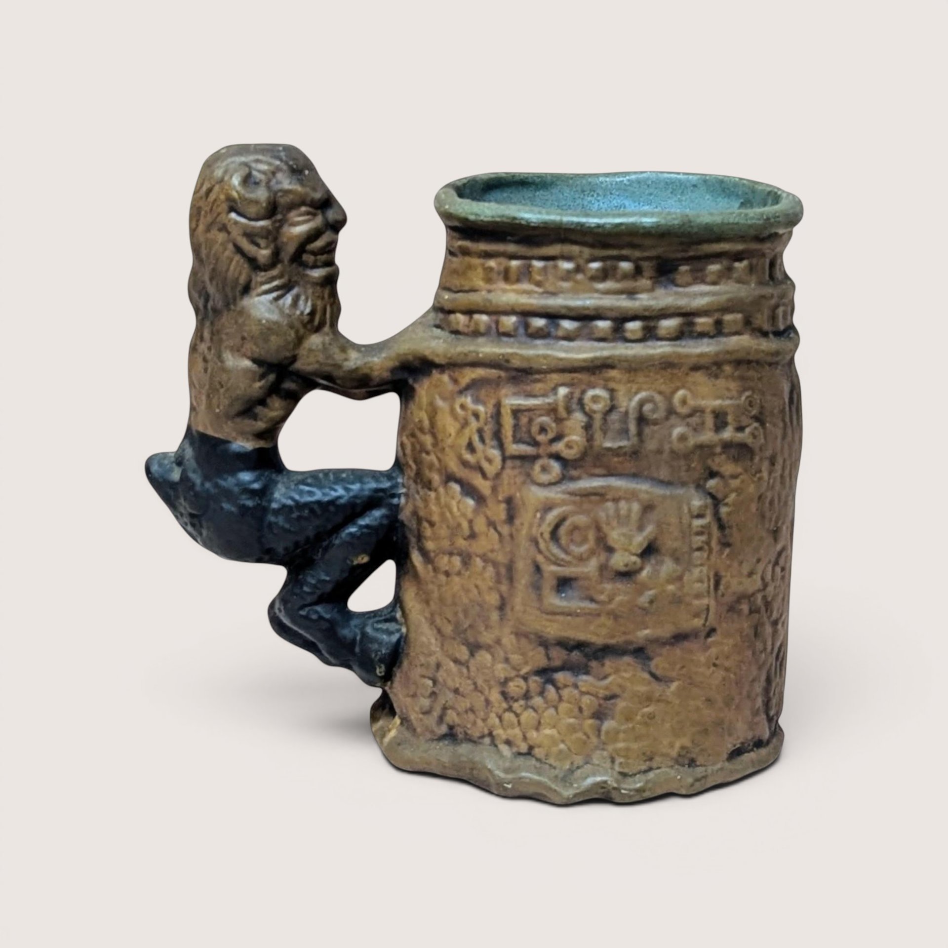 A vintage brown pottery mug featuring a sculpted gargoyle or satyr figure as a handle, centered on a beige background.