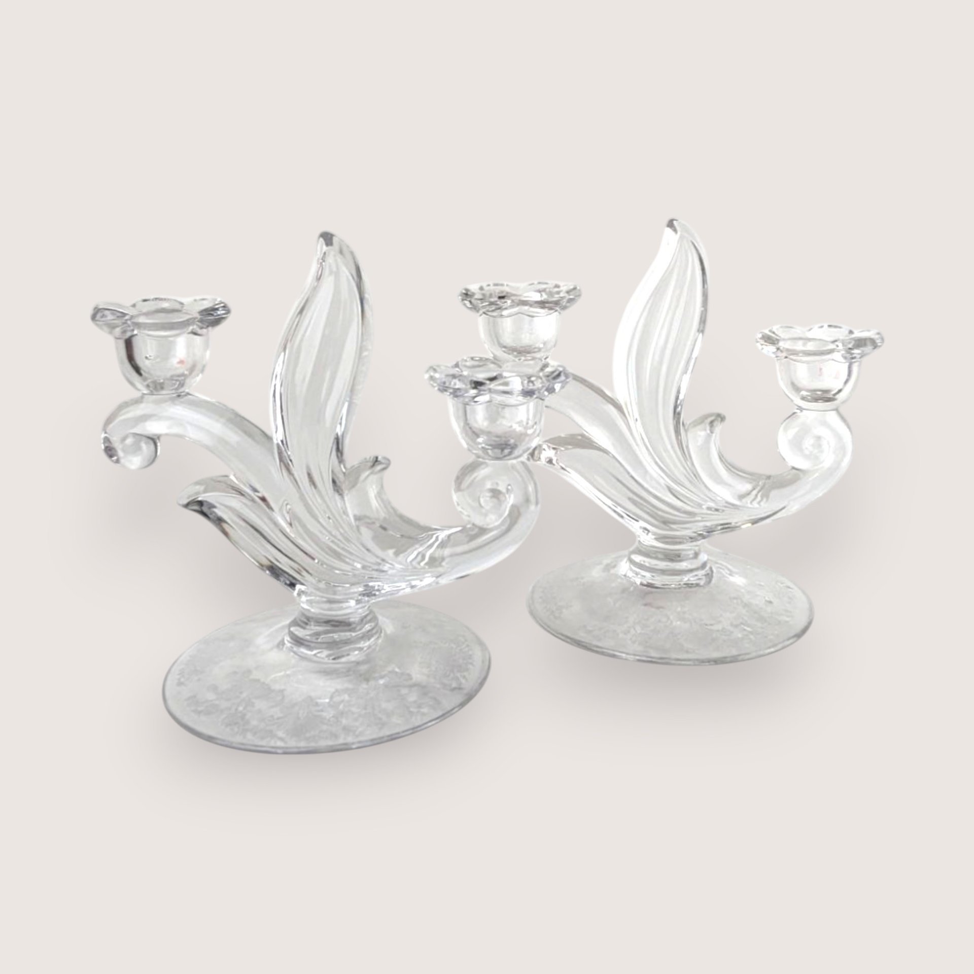 A pair of elegant clear glass candelabras with curved leaf details and flower-shaped cups, centered on a beige background.