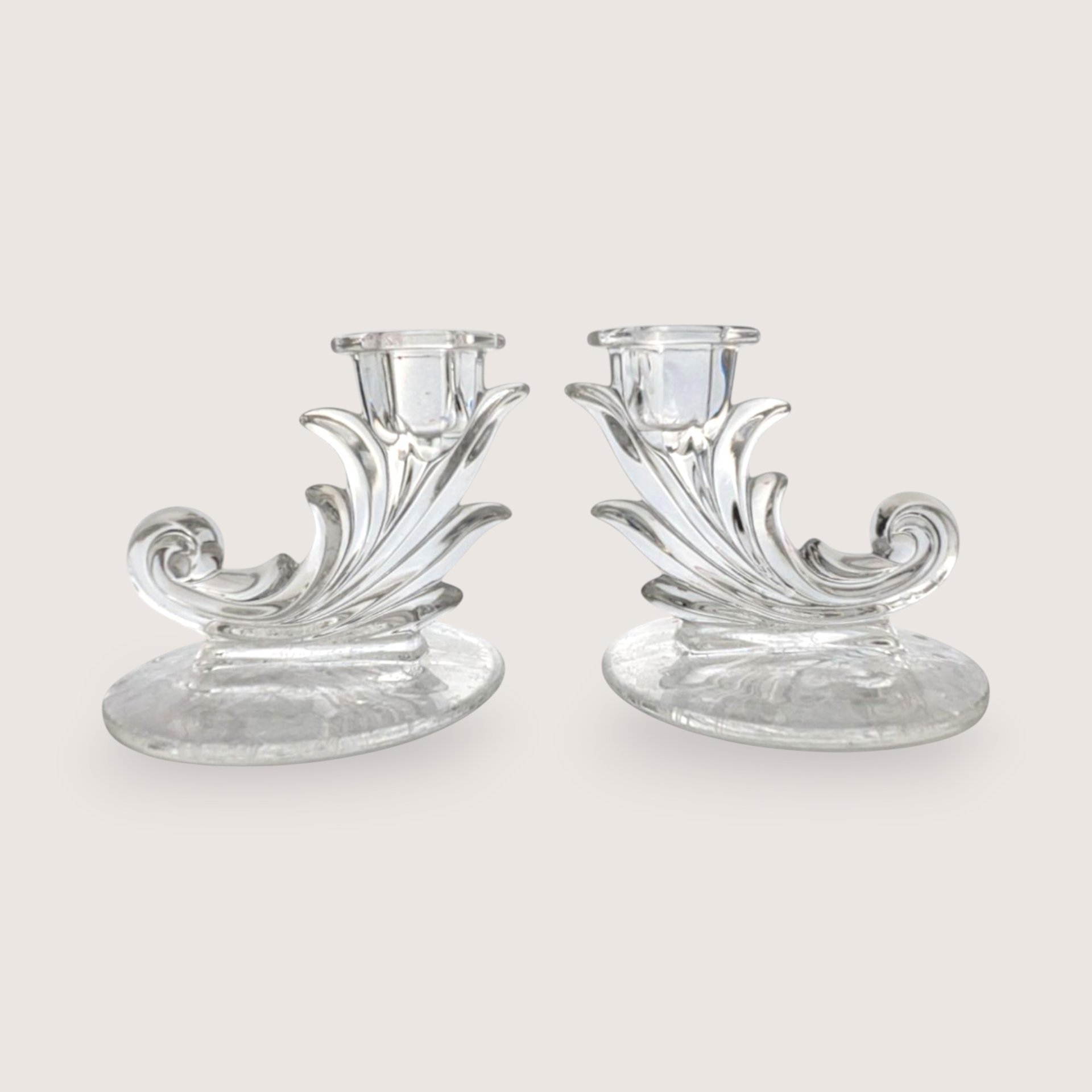 A pair of vintage clear glass cornucopia or leaf-shaped single candlestick holders, centered on a beige background.