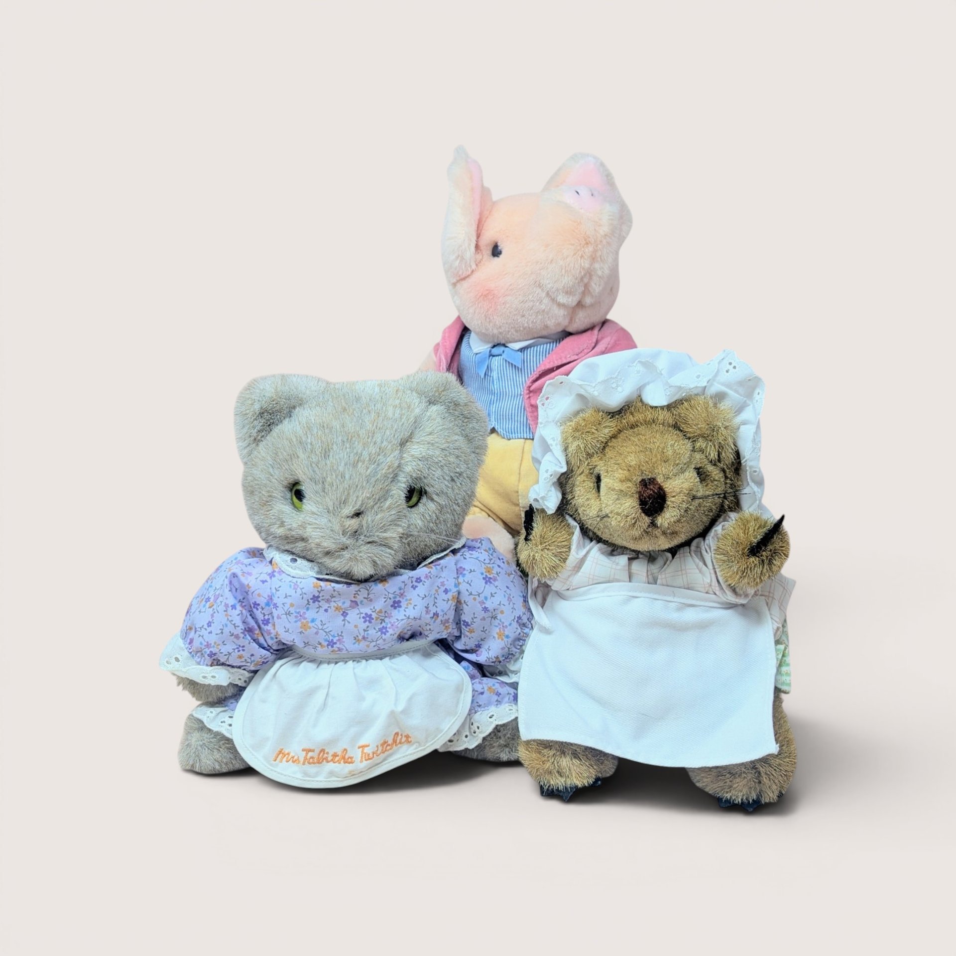 Three Beatrix Potter plush toys, including a grey cat, a hedgehog in a white apron, and a pink pig, on a plain background.