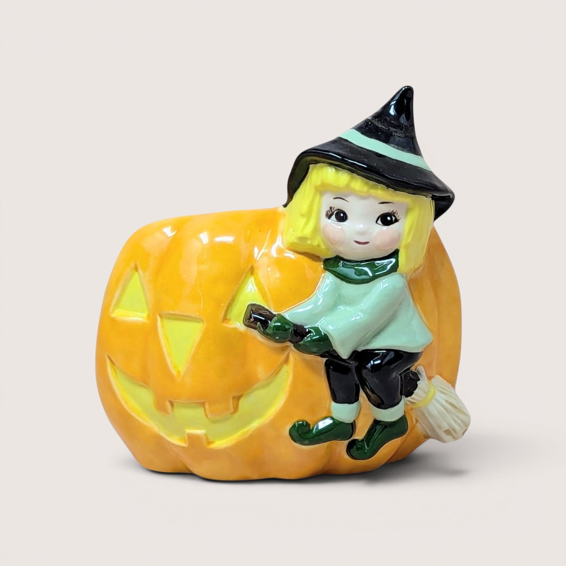 Ceramic Halloween planter featuring a little witch on a broomstick next to a jack-o'-lantern, centered on a plain linen background.
