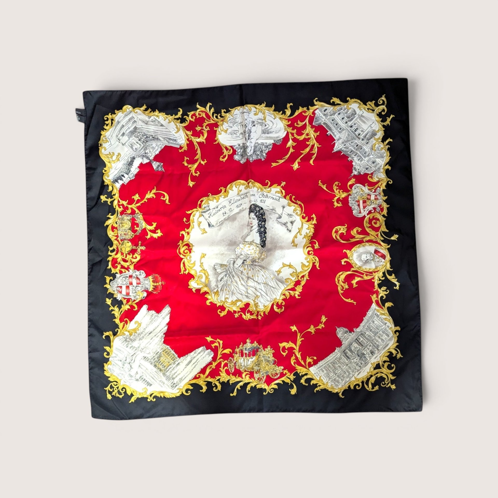 Square scarf featuring a central portrait of Empress Elisabeth of Austria surrounded by illustrated landmarks on a vibrant red background with an ornate gold and black border, centered on a plain linen background.
