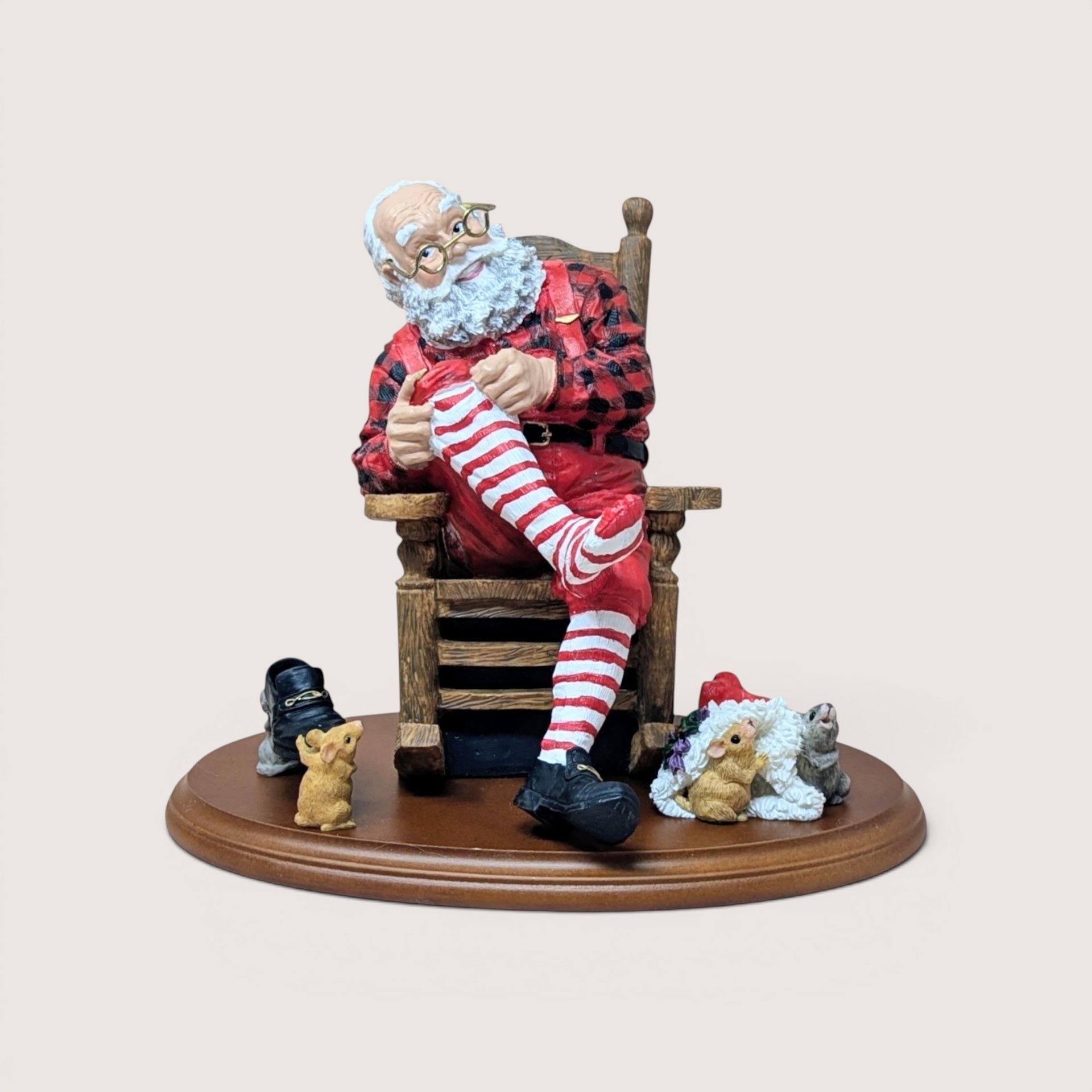 Santa Claus figurine sitting in a rocking chair mending a red and white striped stocking, surrounded by small mice on a wooden base, centered on a plain linen background.