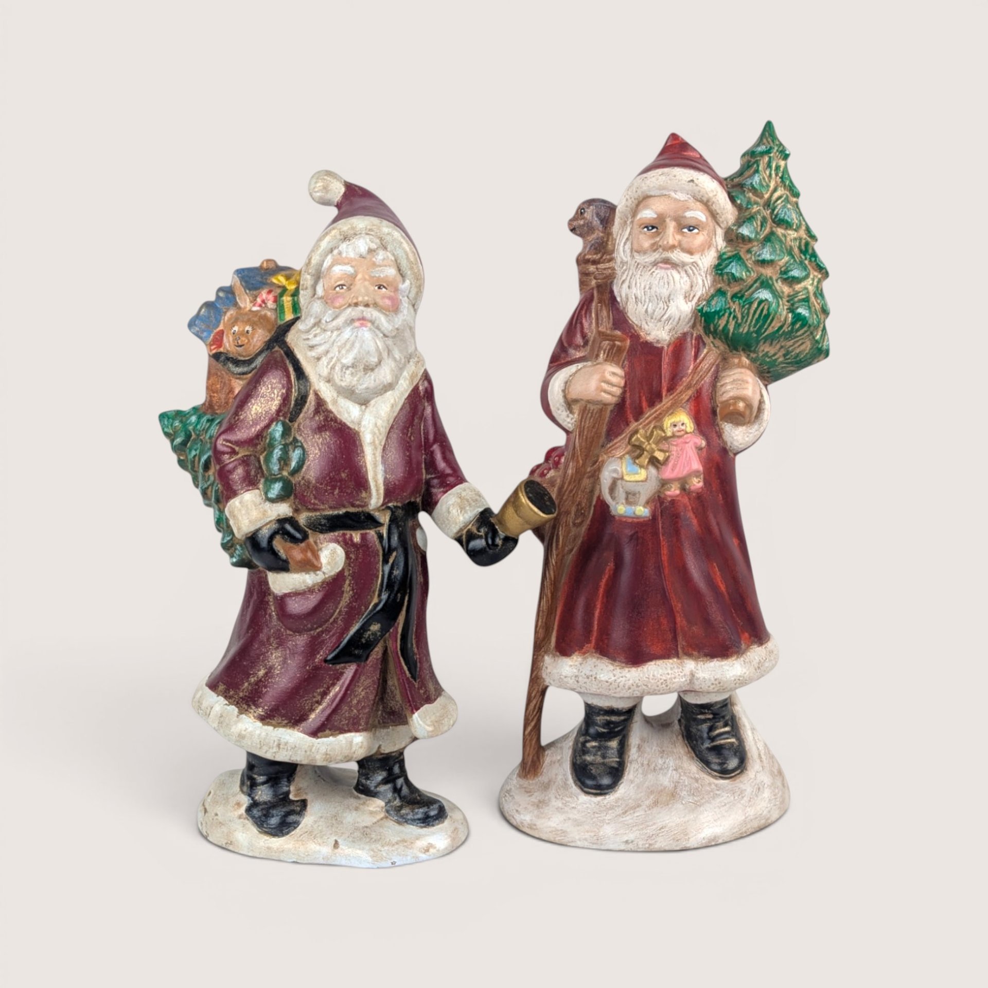 Pair of vintage hand-painted Old World Santa figurines in red coats carrying trees and toys on snowy bases.