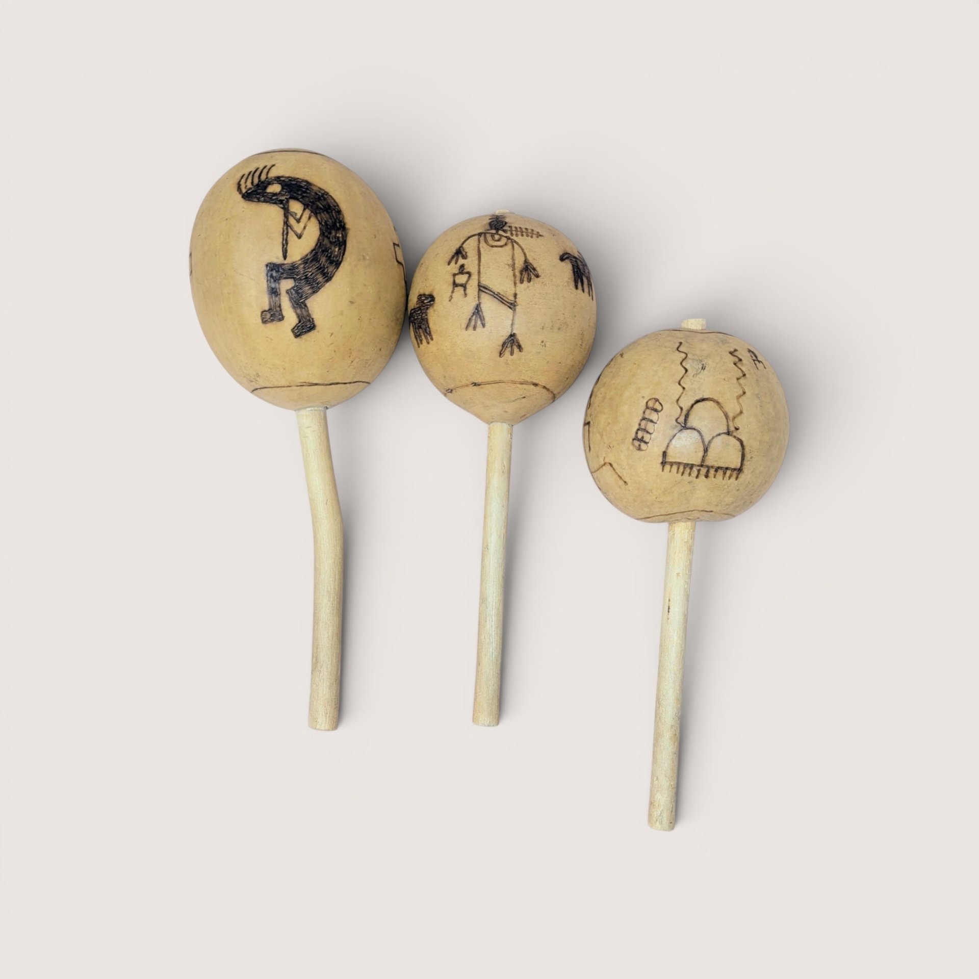 Set of three vintage gourd maracas or rattles with wooden handles, featuring hand-etched Southwestern petroglyph and Kokopelli designs, centered on a plain linen background.