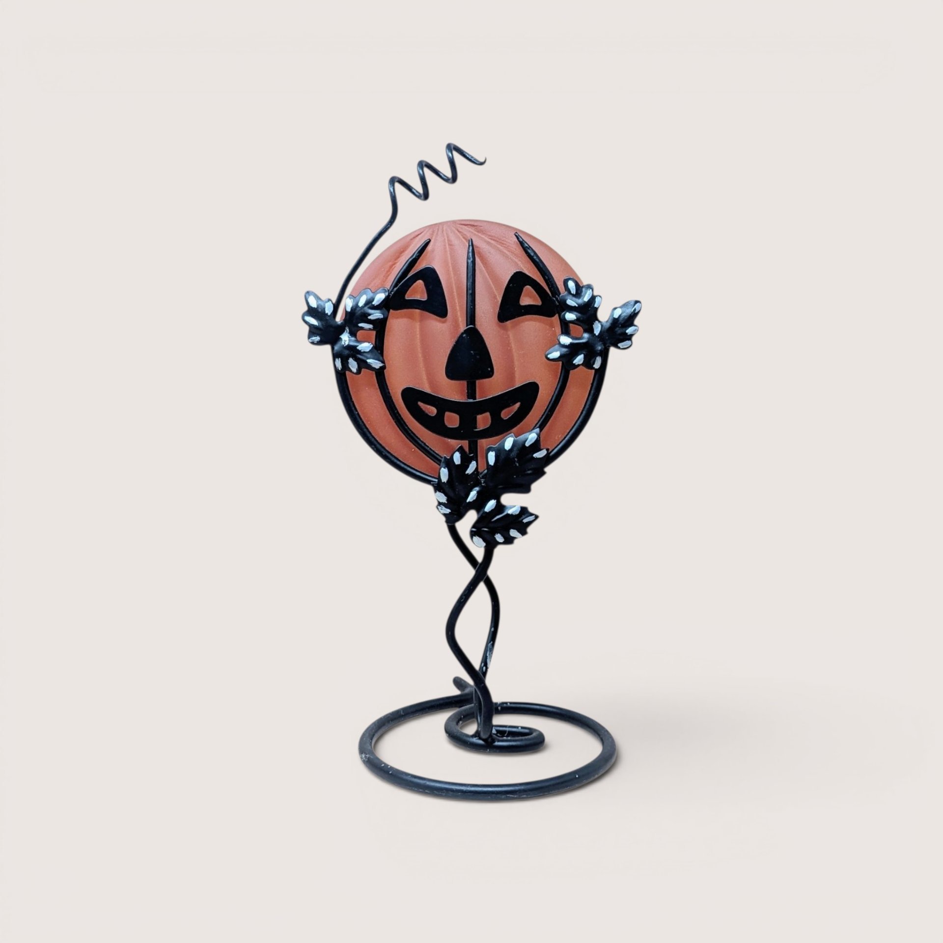 Vintage frosted orange glass jack-o'-lantern face insert sitting inside a black metal wire frame stand accented with leaves and a curly vine top, centered on a plain linen background.