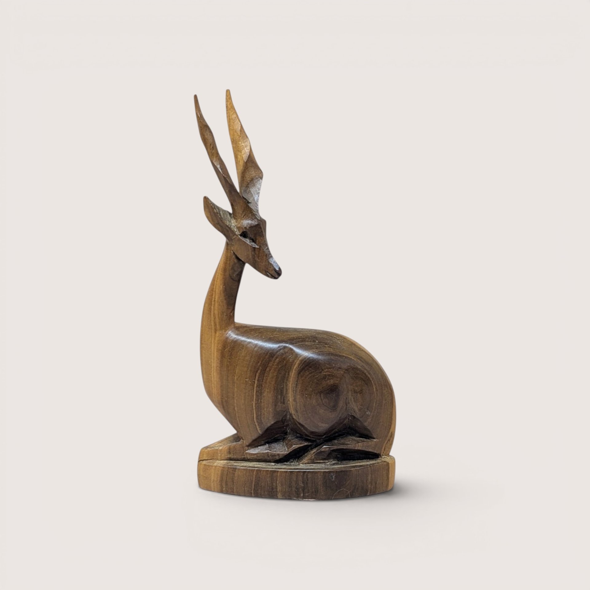 Hand-carved wooden gazelle or antelope figurine depicted in a recumbent, bedded position on an oval base, featuring prominent twisted horns and a rich, polished natural wood grain finish, centered on a plain linen background.