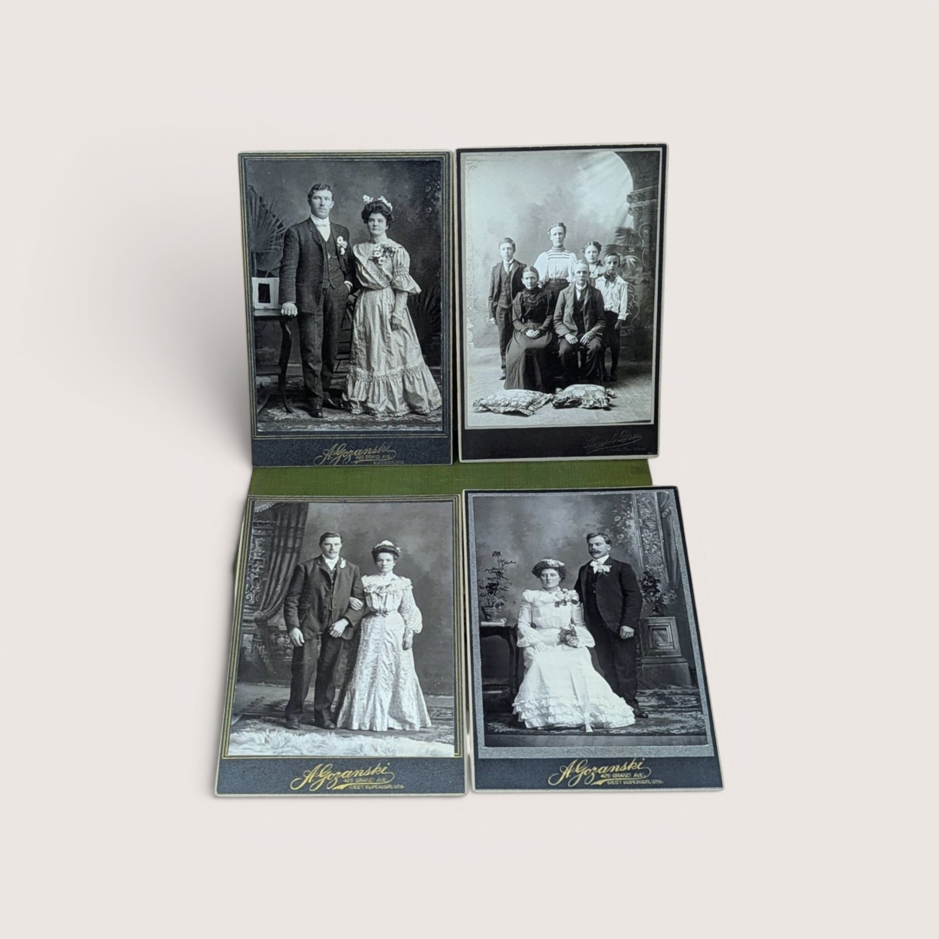 Collection of four vintage black-and-white cabinet card photographs depicting formal family and couple portraits from the late 19th or early 20th century, arranged in a two-by-two grid on a green fabric surface, centered on a plain linen background.