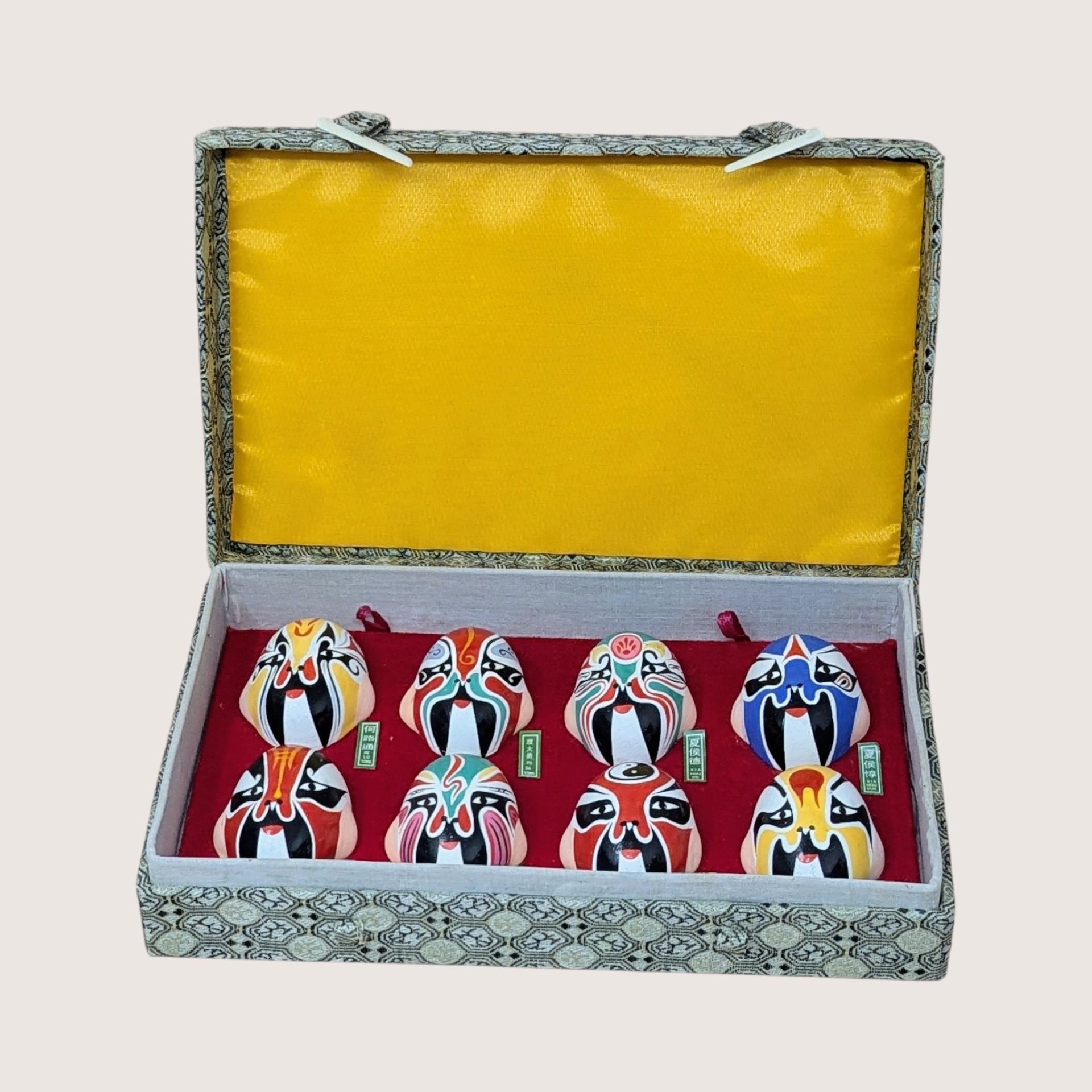 A vintage boxed display of eight hand painted Beijing Opera clay masks resting on a soft pink background. The vibrant, colorful theatrical facial makeup designs are arranged on red velvet inside a fabric covered case with a bright yellow satin lined lid.