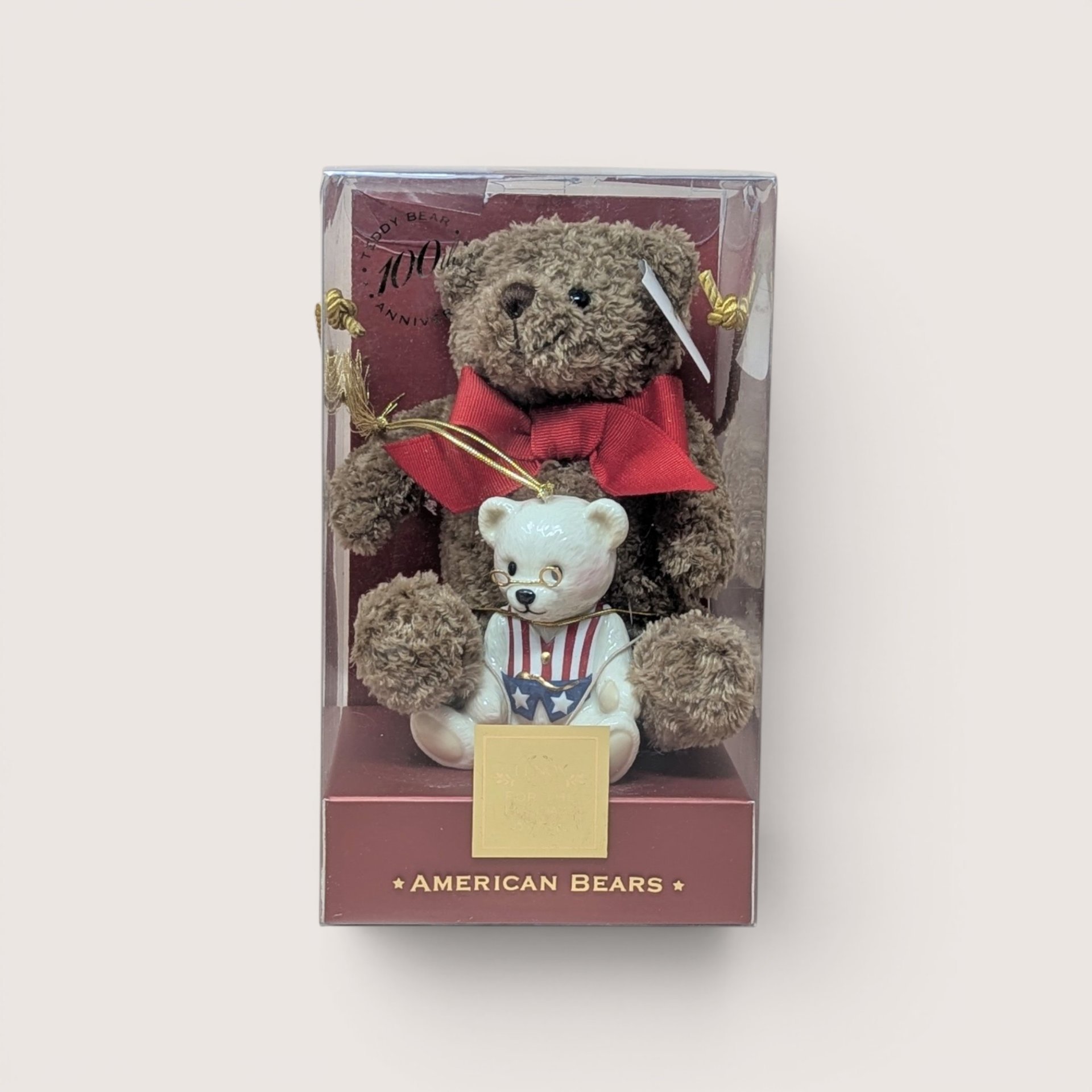 Boxed collector's set titled "AMERICAN BEARS" celebrating the Teddy Bear 100th Anniversary, containing a small brown plush teddy bear wearing a large red bow paired with a white porcelain teddy bear figurine dressed in a patriotic red, white, and blue vest with wire-rimmed glasses, centered on a plain linen background.