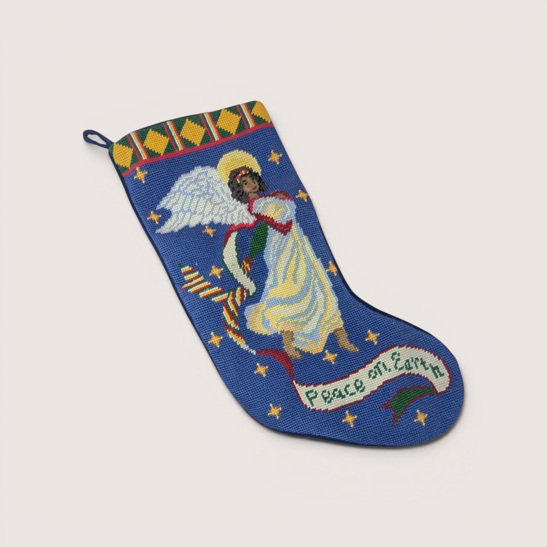 Vintage needlepoint Christmas stocking featuring a winged angel with a gold halo holding a flowing banner that reads "Peace on Earth" across a blue, star-accented background with a geometric diamond pattern along the top cuff, centered on a plain linen background.