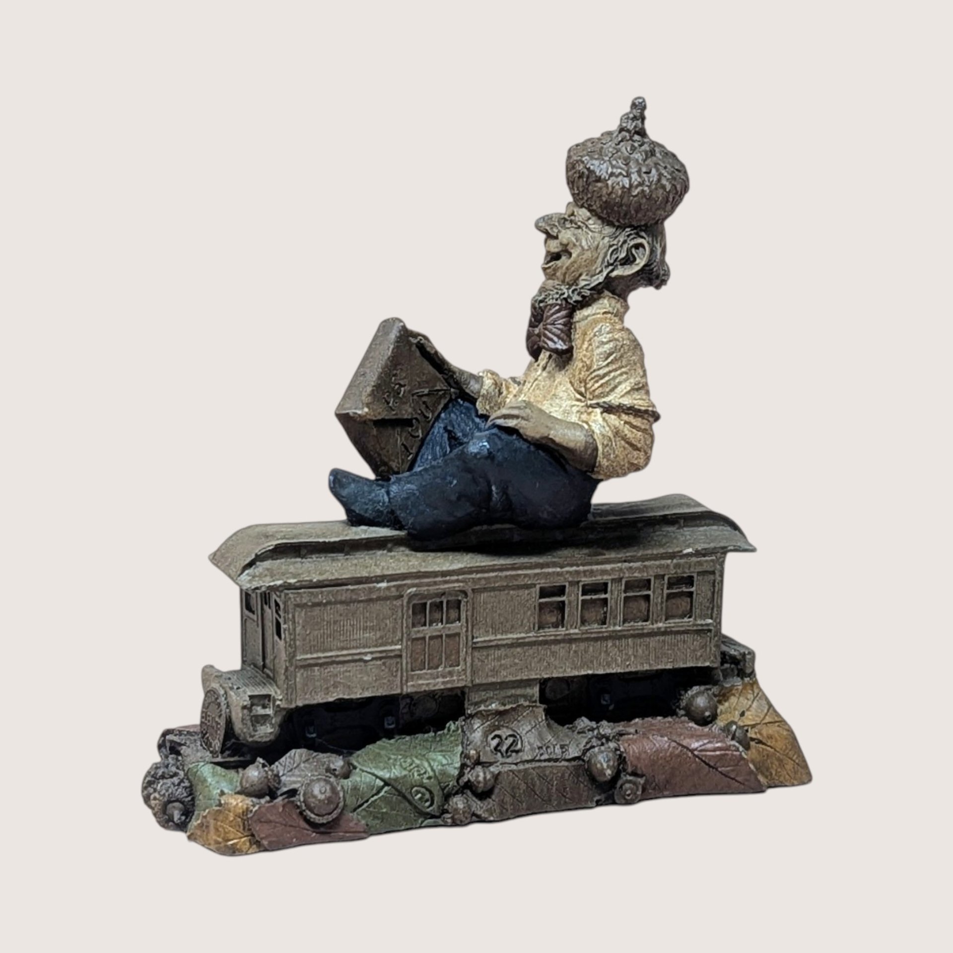Vintage Tom Clark Cairn studio gnome figurine depicting a gnome named "Cole" wearing an acorn-cap hat, seated on top of a miniature trolley car or train passenger car numbered 22, resting on a base of textured leaves and acorns, centered on a plain linen background.